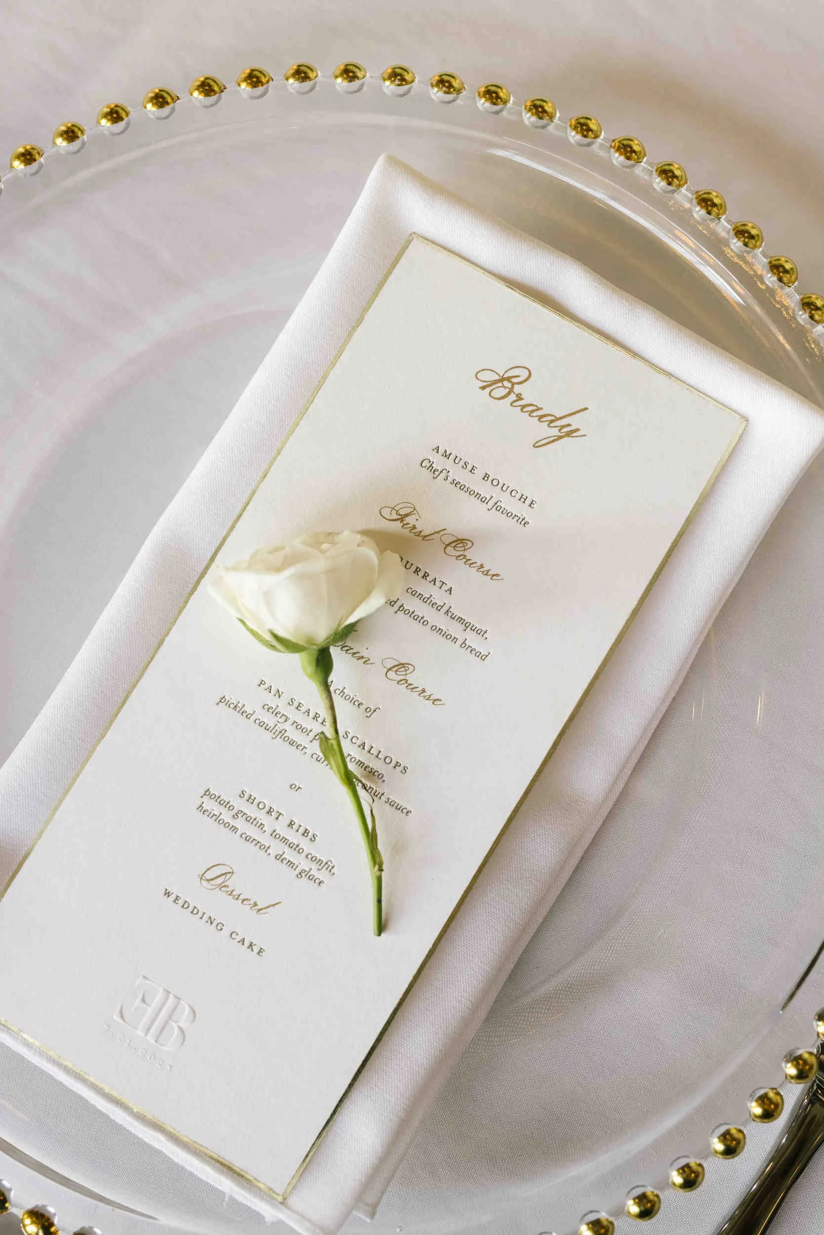 Elegant white place setting with a gold-embossed wedding cake menu card on a white napkin, a white flower, and a decorative glass plate with gold beads around the edge.