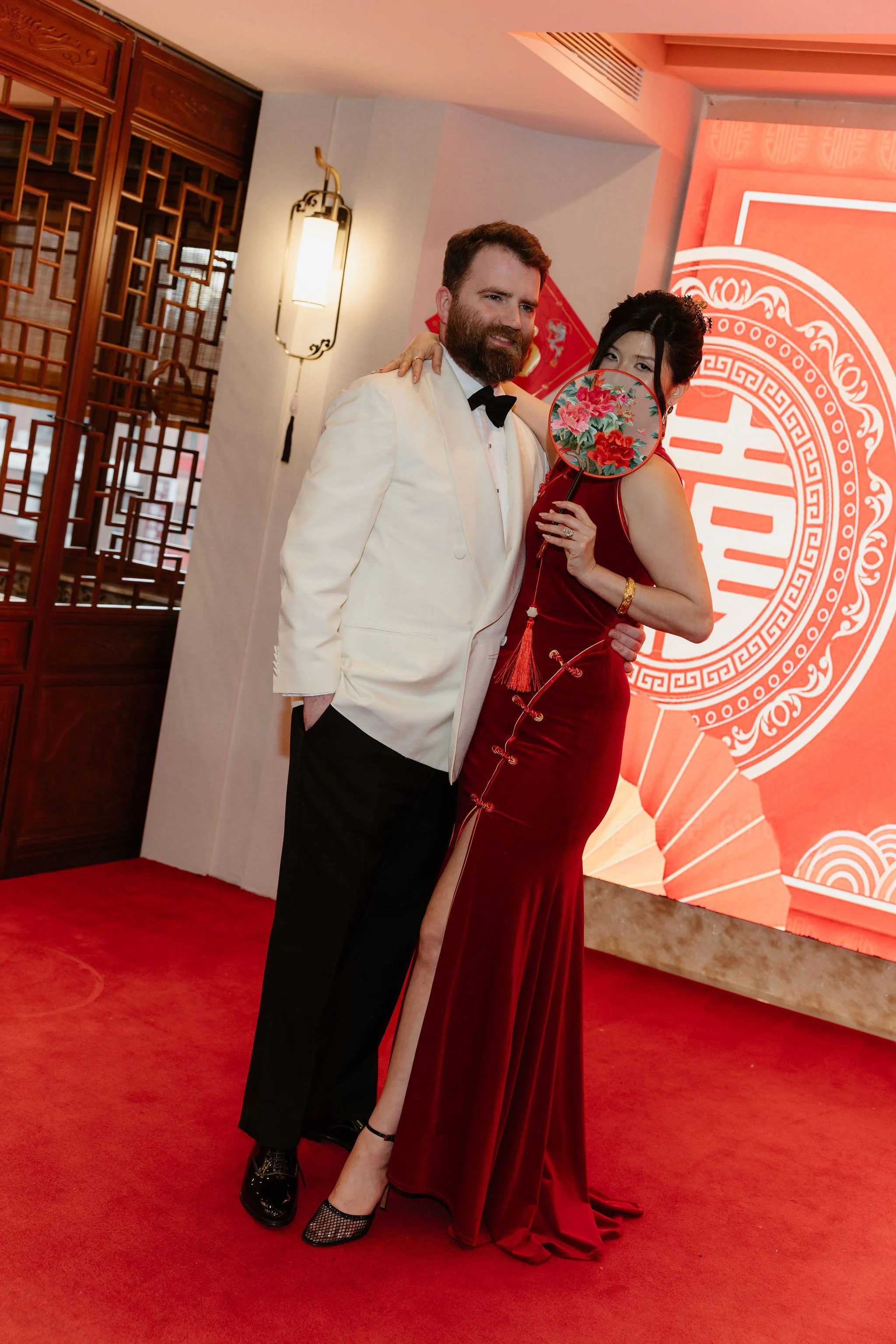 A man with a beard in a white tuxedo and a woman in a red dress with a lace-up design hold a fan with pink peach blossoms. They stand on a red carpet in front of a large red Chinese-themed backdrop featuring the double happiness symbol.
