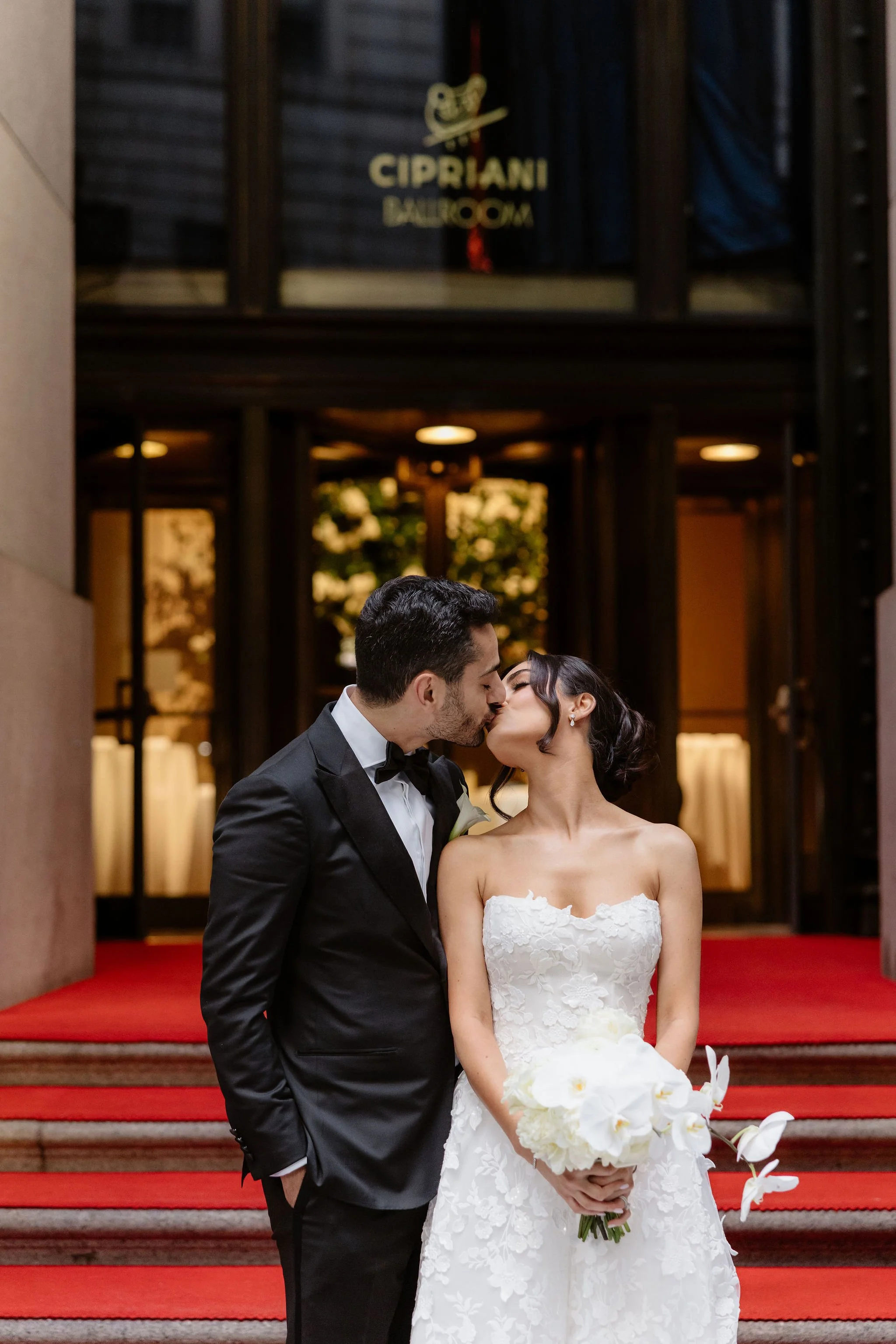 A newlywed couple sharing a kiss on the steps of a building labeled Cipriani Ballroom. The groom is dressed in a black tuxedo and the bride in a strapless white wedding dress holding a bouquet of white flowers.