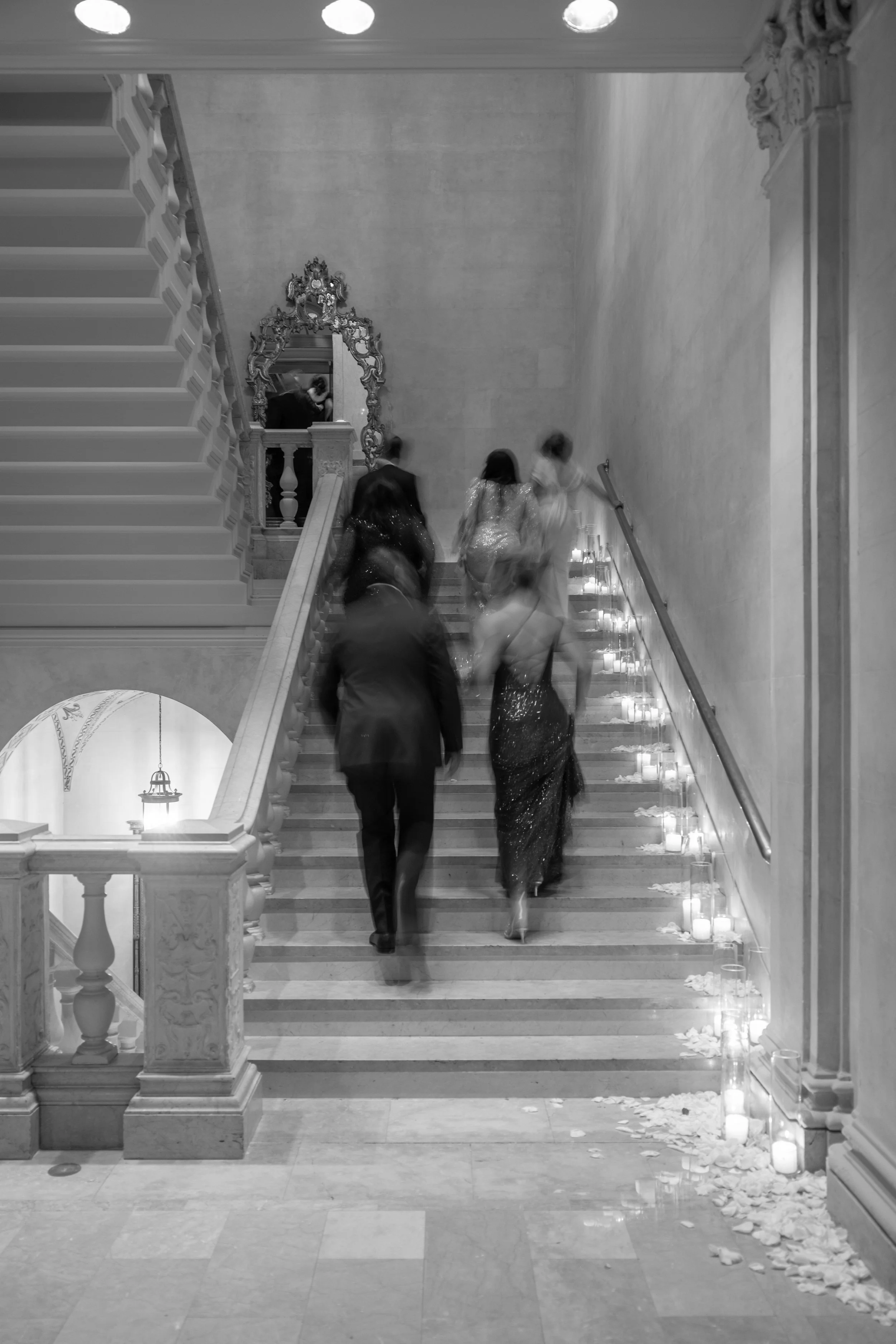 A black and white photo of a staircase at a formal event, with a few women in evening gowns ascending. Candles and scattered rose petals are along the side of the stairs, and an ornate mirror hangs above.