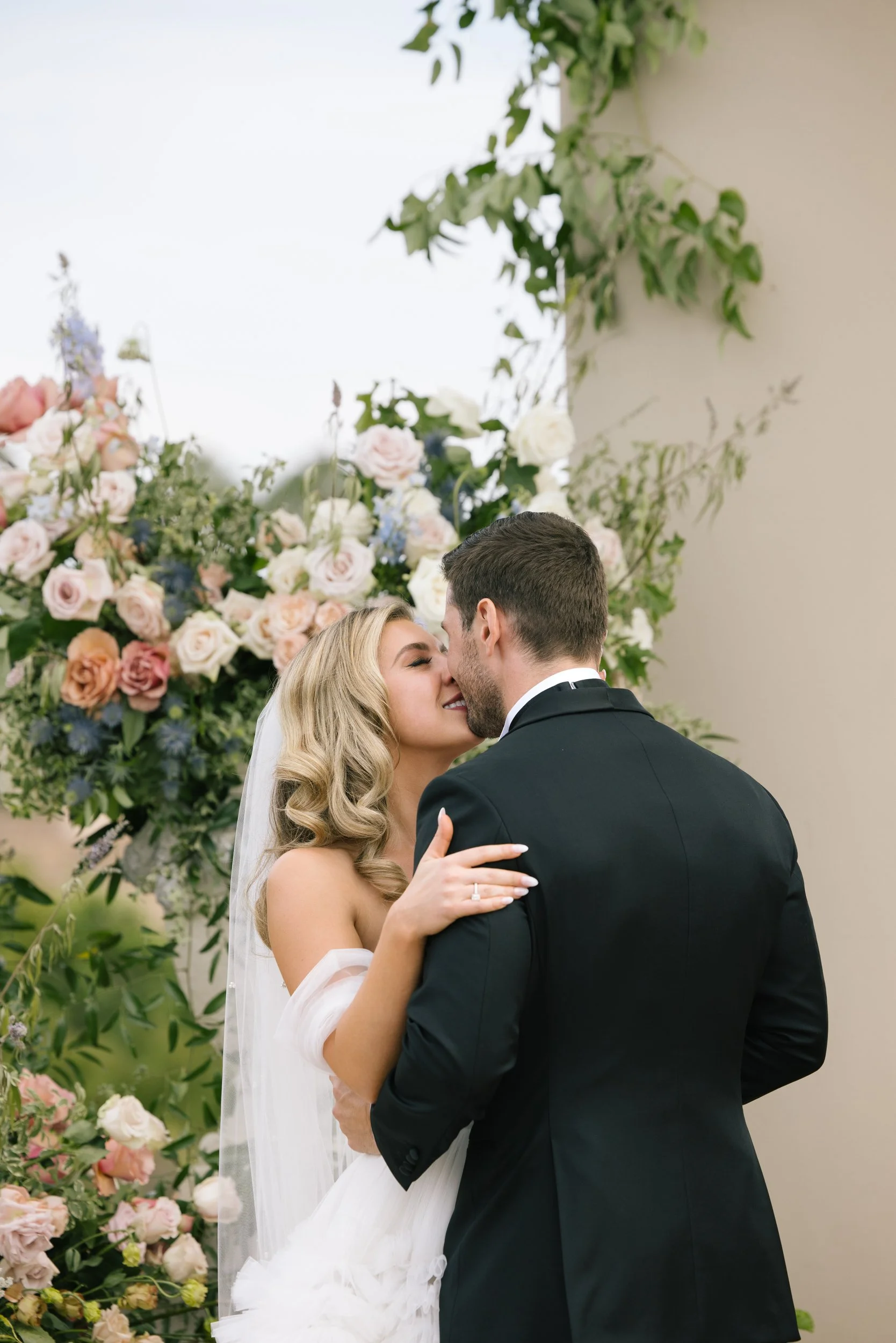 A bride and groom sharing a kiss during their wedding ceremony, with a background of pink and white flowers and green foliage.
