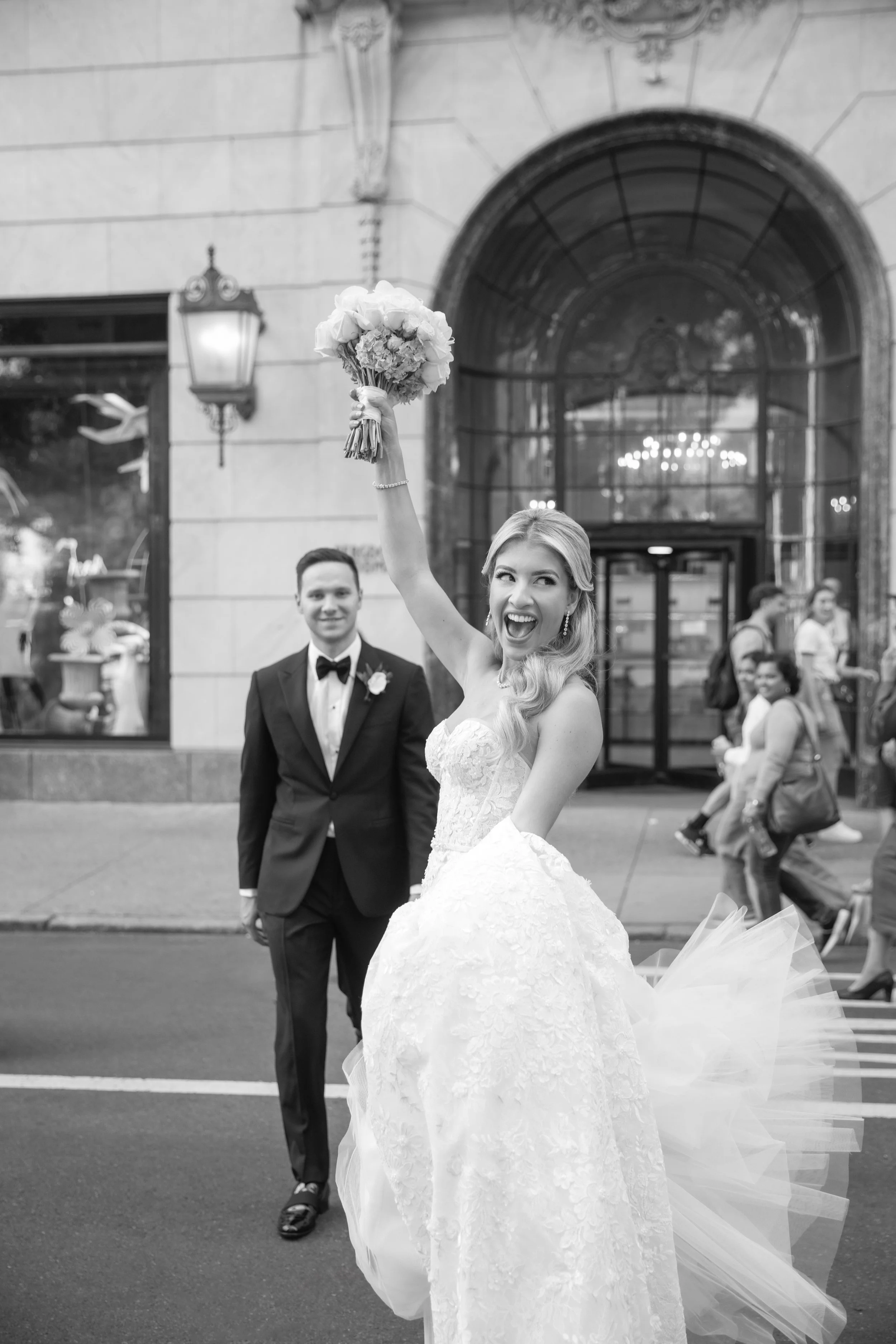 A bride holding a bouquet high in the air, smiling, with the groom standing behind her in front of a building with large windows and people in the background.