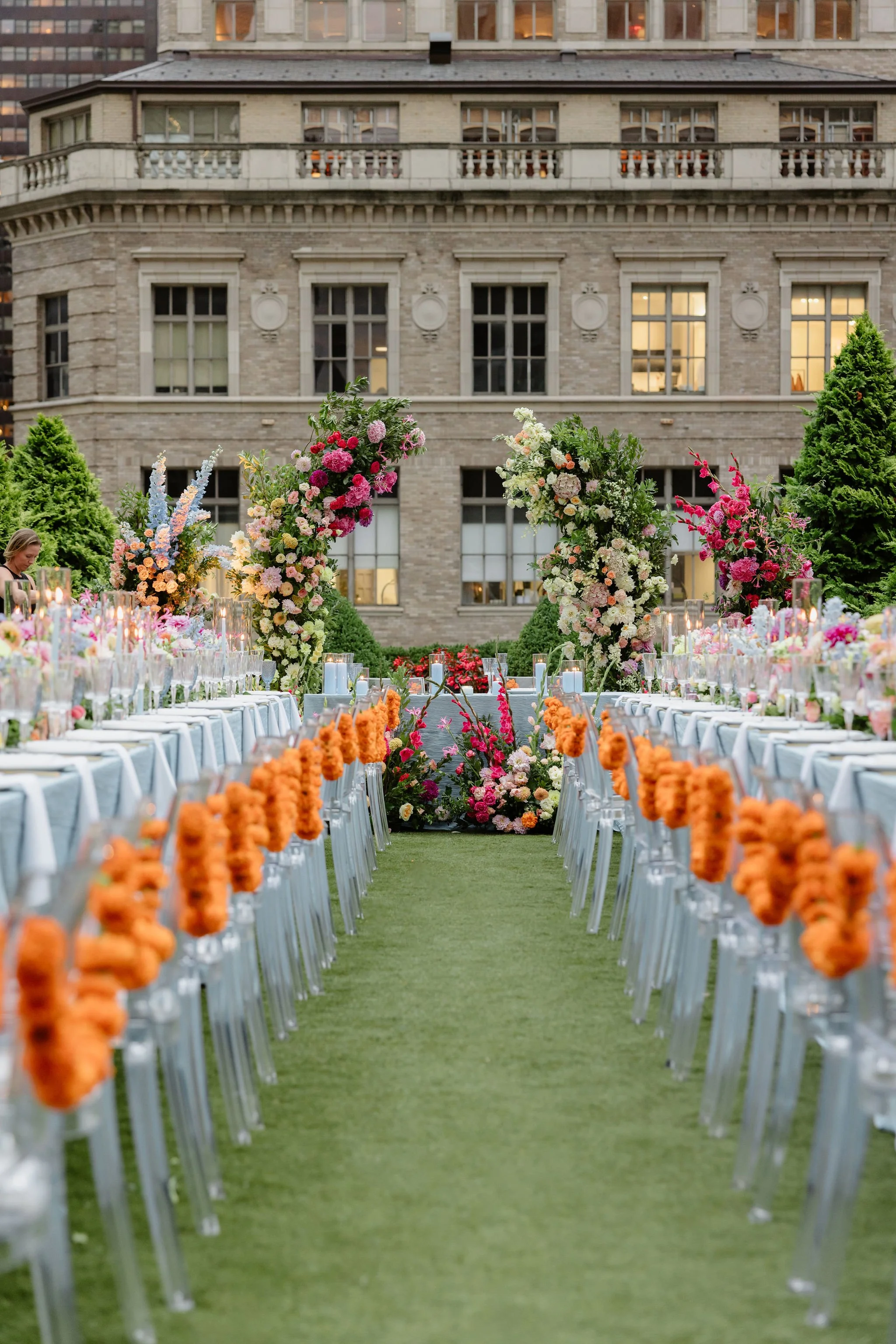 Outdoor event setup with long tables decorated with orange flowers in vertical garlands, surrounded by floral arrangements and greenery, with a brick building in the background.