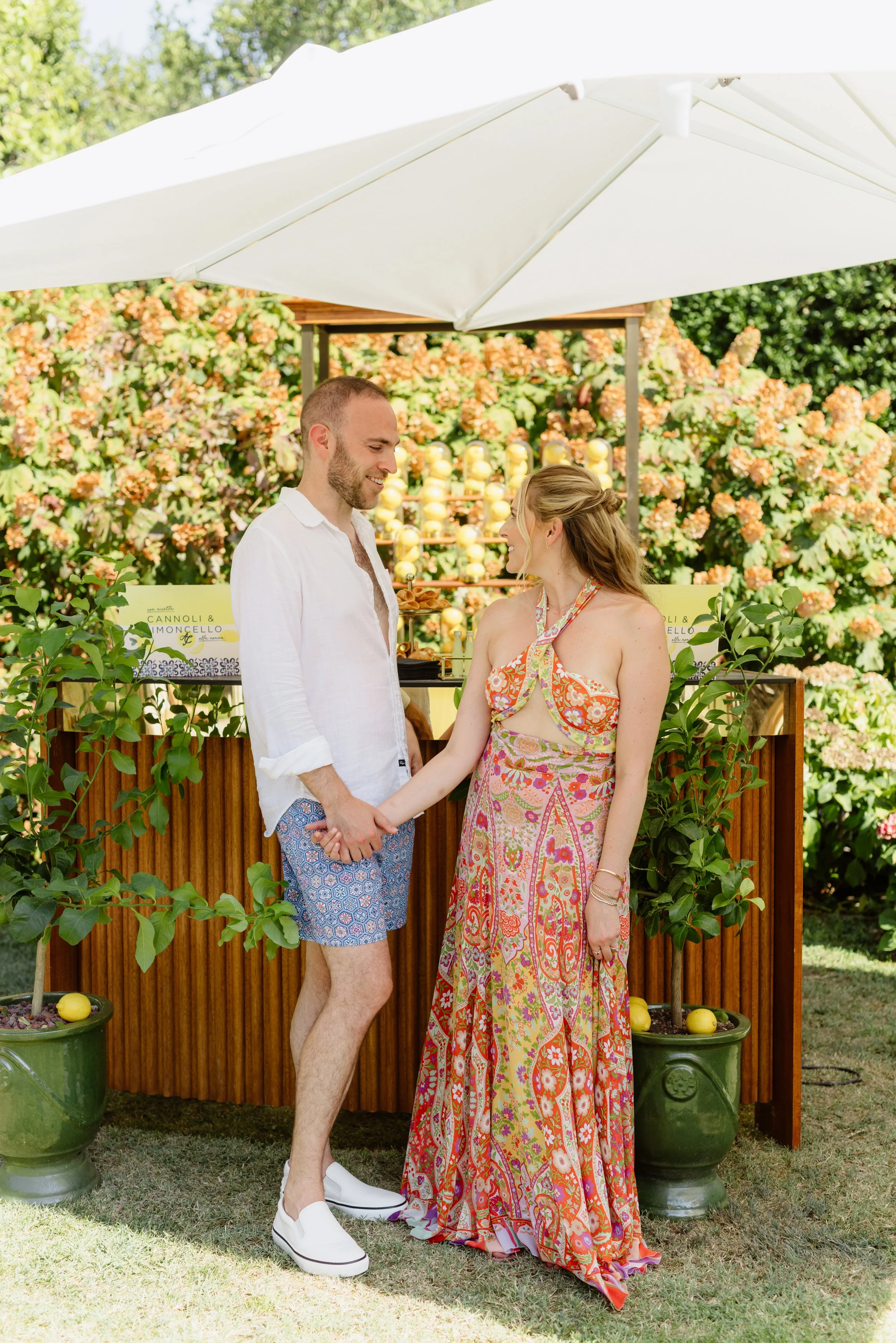 A man and woman holding hands, smiling at each other, standing outdoors under a large white umbrella. The woman is wearing a colorful, patterned dress, and the man is wearing a white shirt and patterned shorts. There are potted lemon trees and a wood