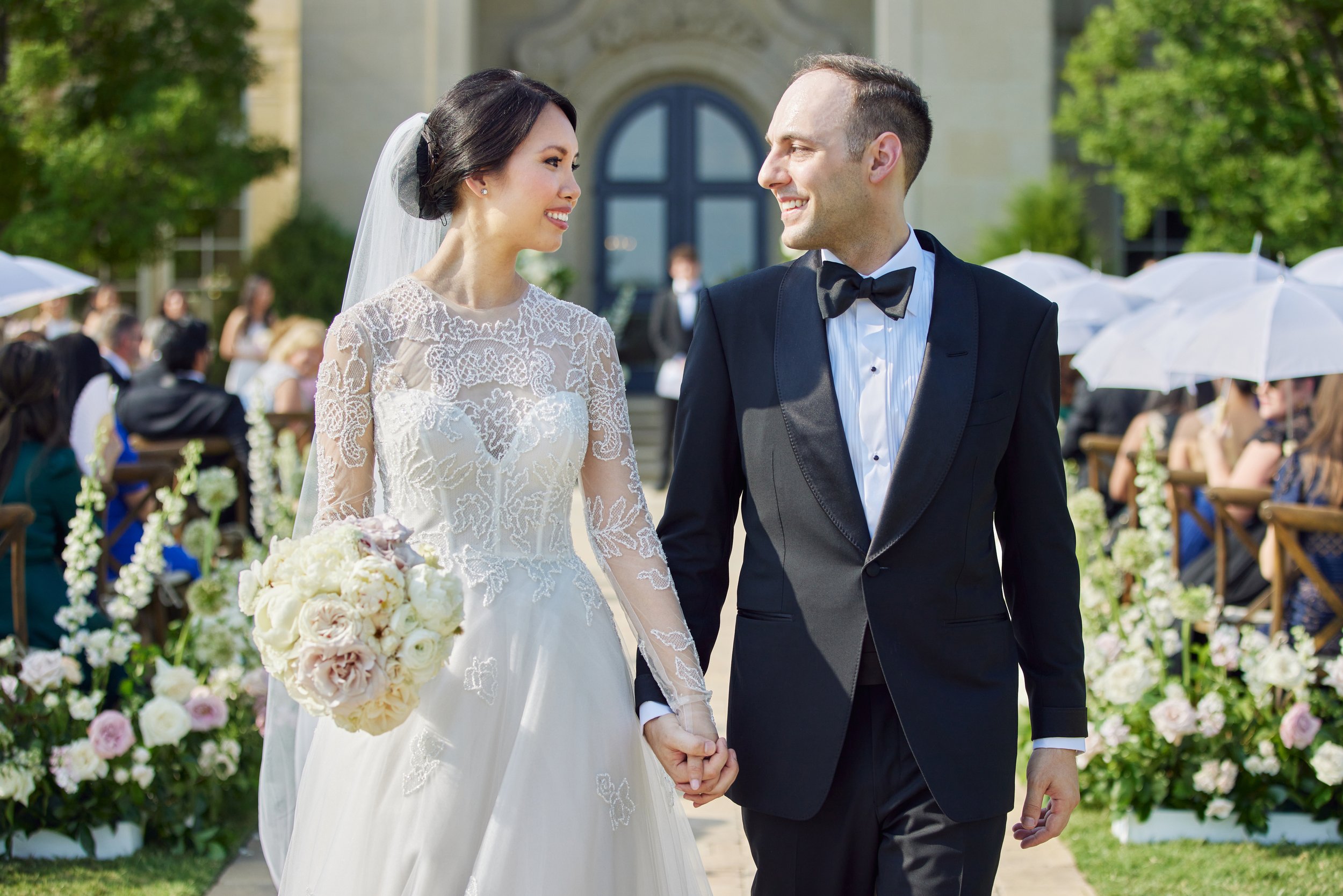 A newlywed couple holding hands and smiling at each other during their outdoor wedding ceremony, with guests and floral decorations in the background.