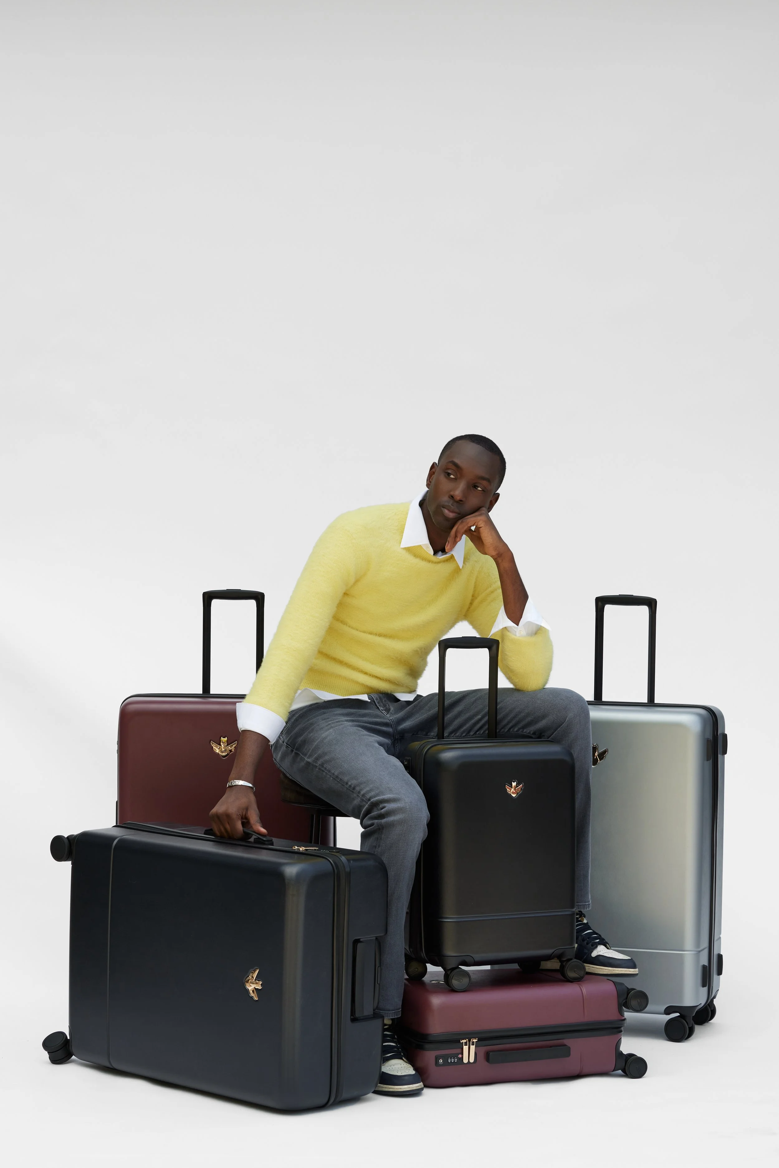 A young man sitting on stacked suitcases with his head resting on his hand, surrounded by additional suitcases, all against a plain white background.
