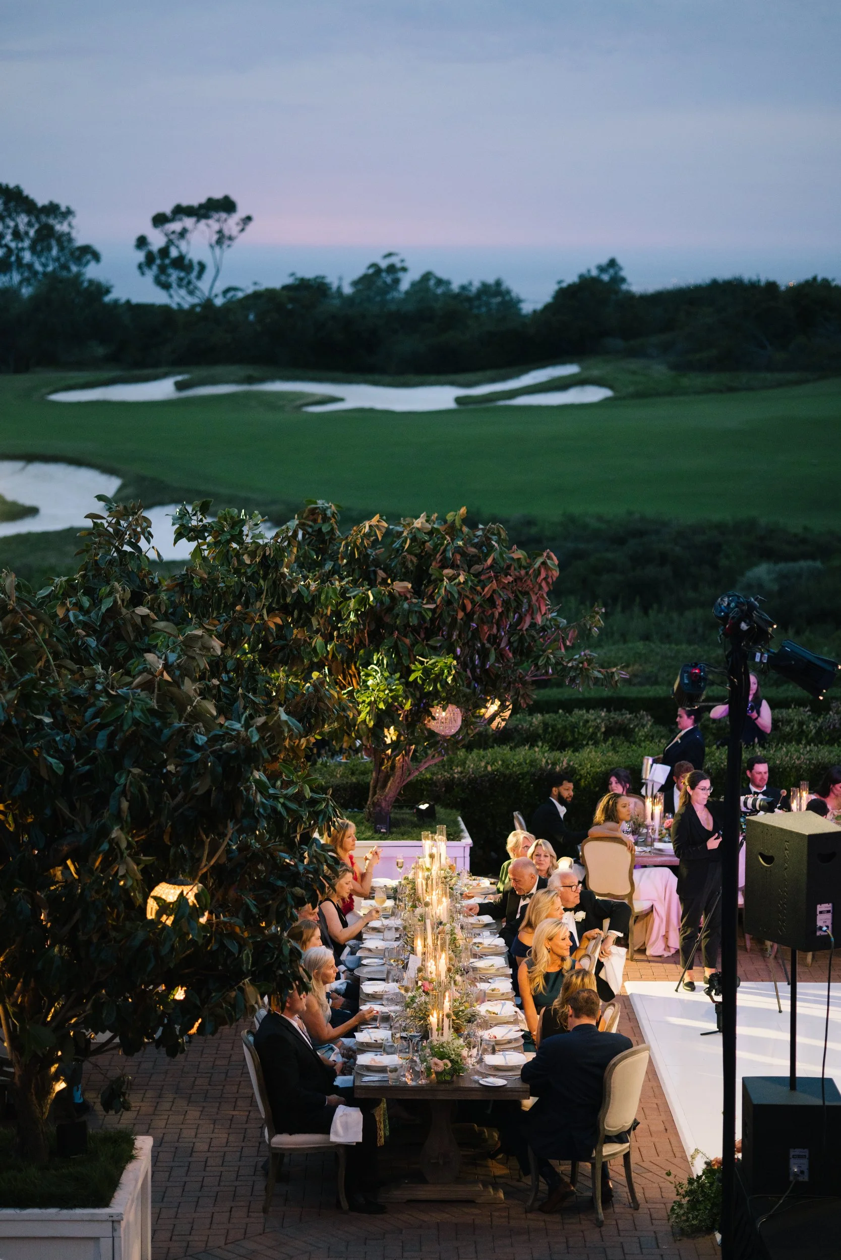 An outdoor formal dinner event with guests seated at long tables decorated with candles and floral centerpieces, overlooking a golf course with trees in the background during dusk.