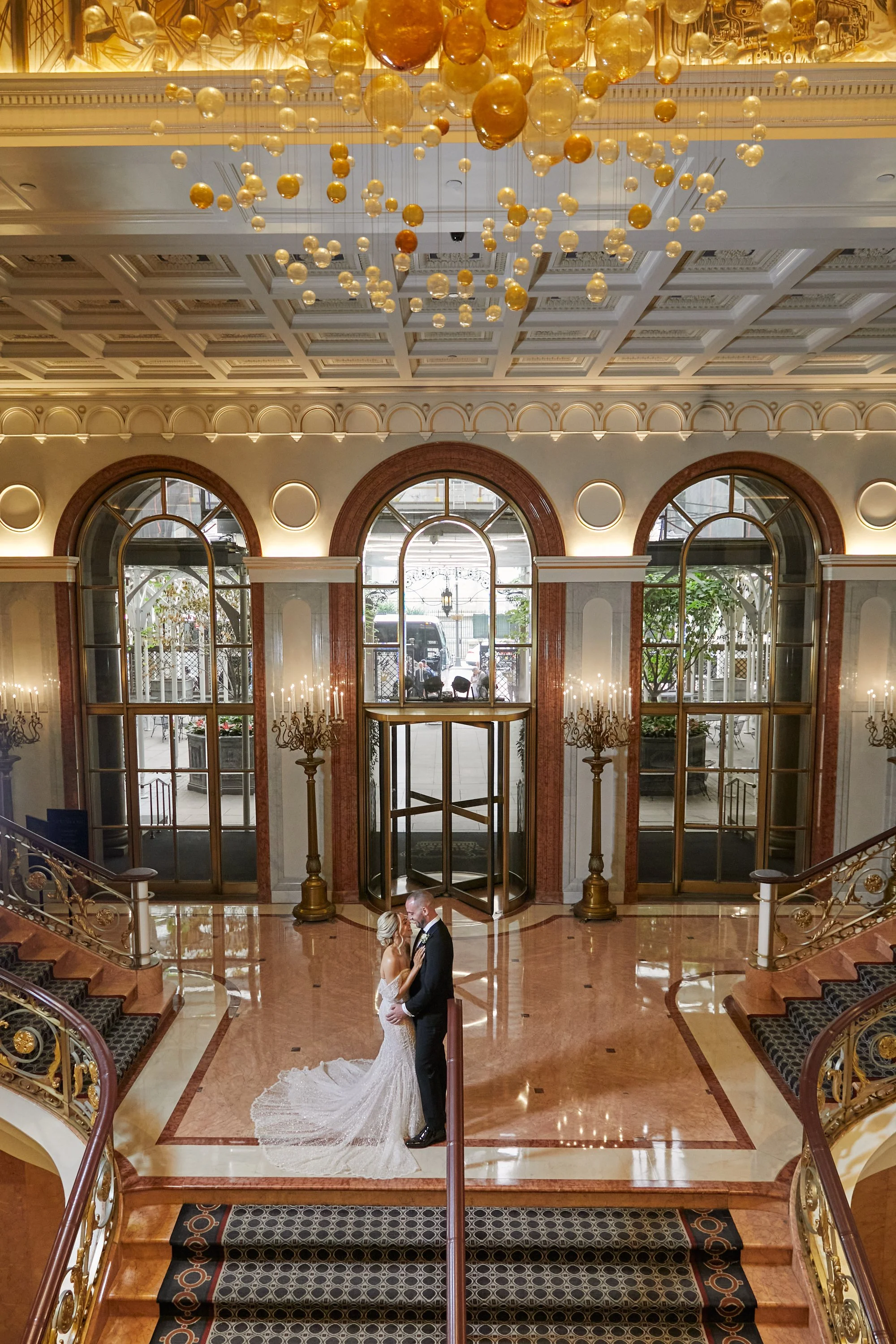 A bride and groom are dancing in a luxurious, elegant hotel lobby with high ceilings, large arched windows, ornate chandeliers, and intricate gold and wood detailing.