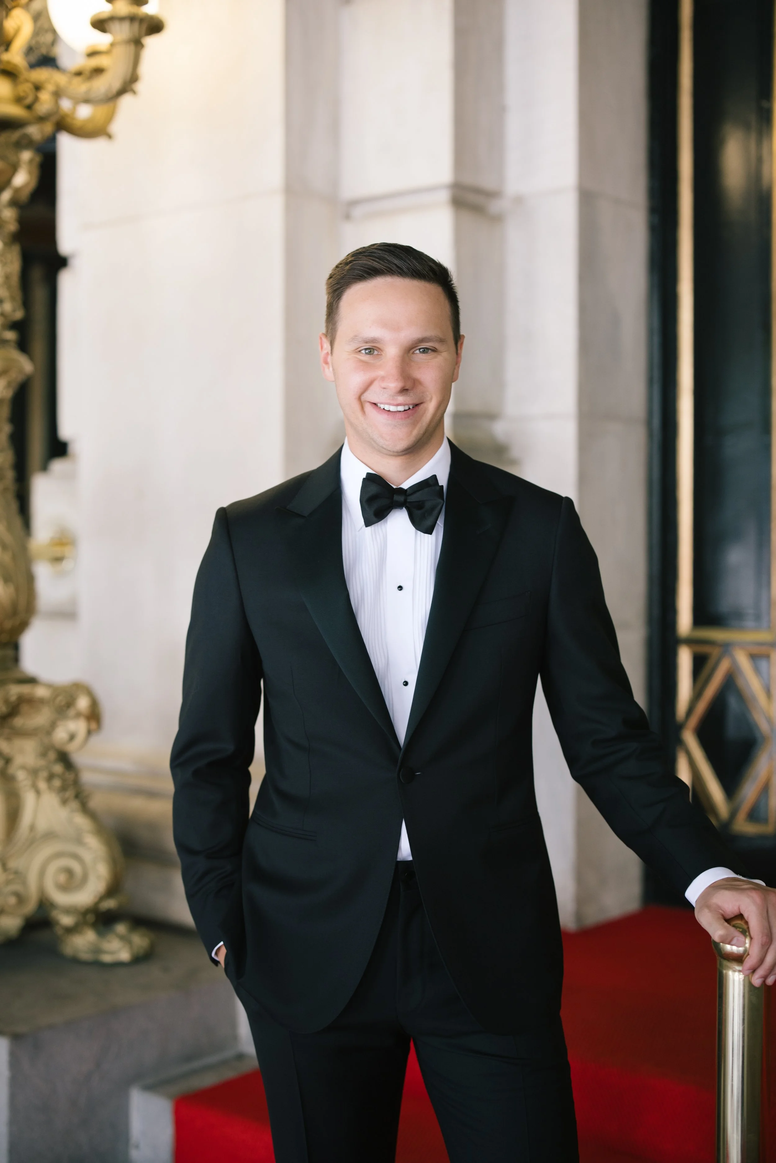 A young man dressed in a black tuxedo with a bow tie, smiling, standing on a red carpet in an elegant interior with ornate decor.