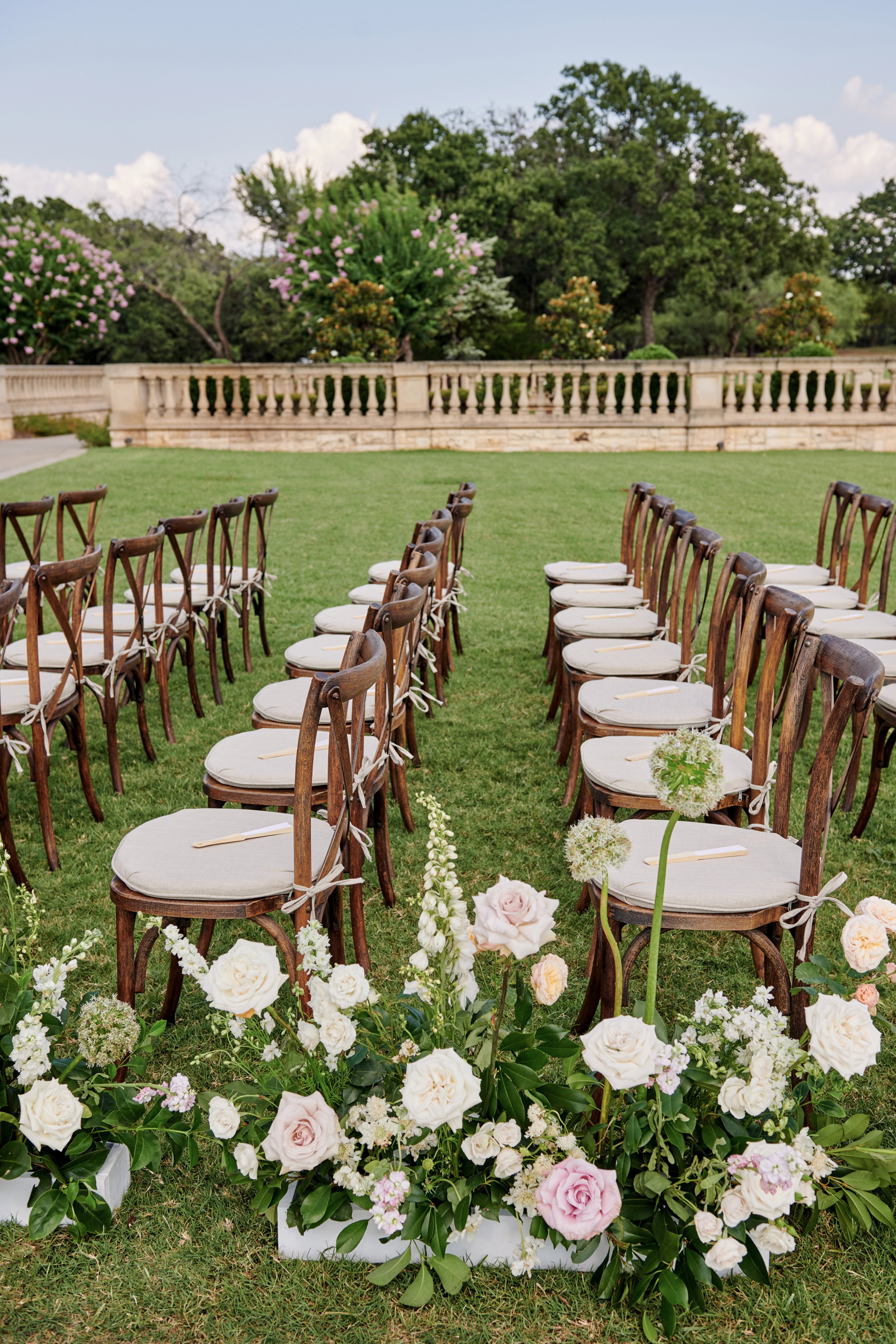 Outdoor wedding setup with rows of wooden chairs with white cushions facing a floral arrangement of pink and white roses and other flowers, surrounded by greenery and trees under a partly cloudy sky.