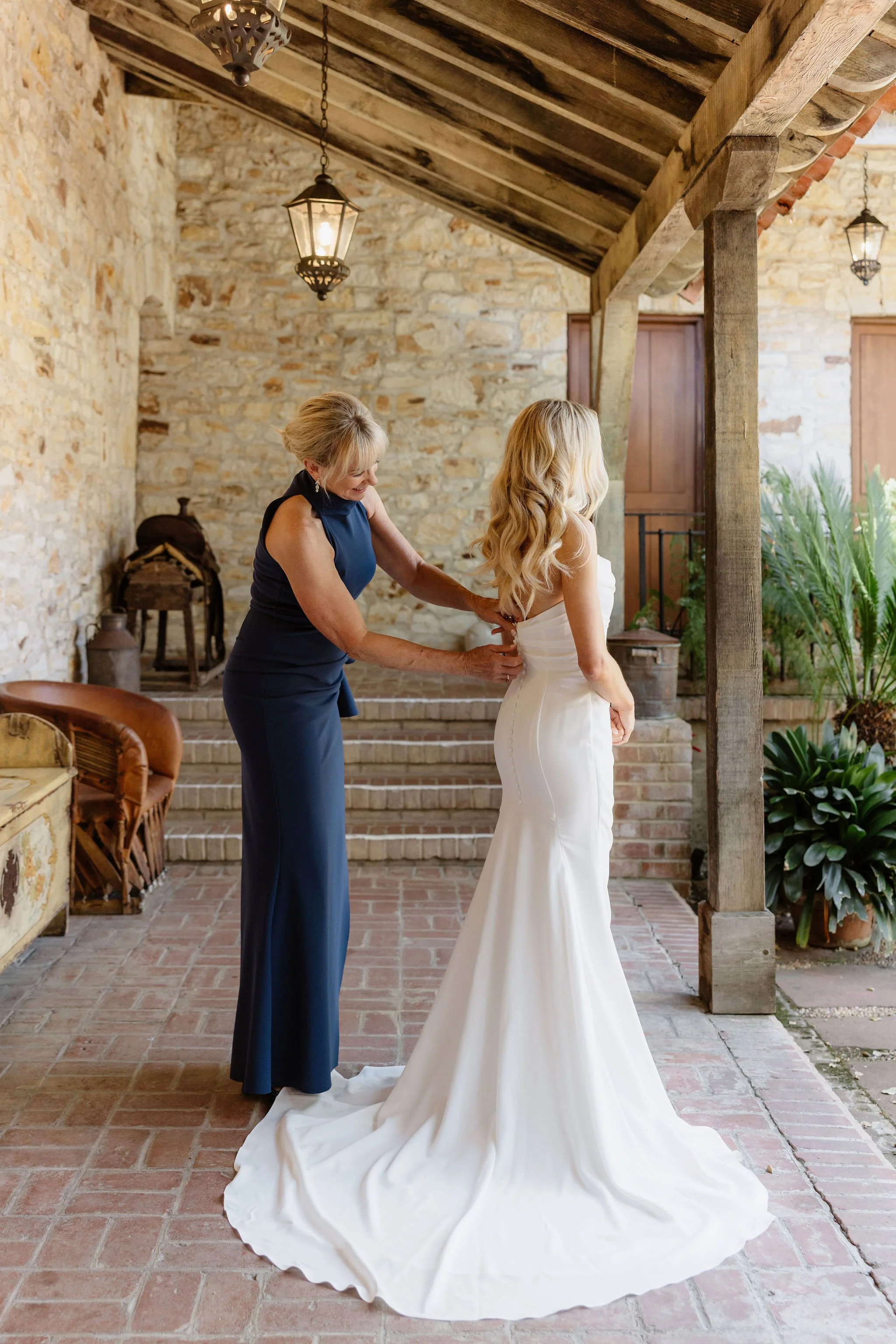 A woman in a strapless white wedding dress is being helped with her dress by another woman in a dark blue dress. They are standing on a brick patio under a wooden pergola with lanterns, next to a stone wall and lush green plants.