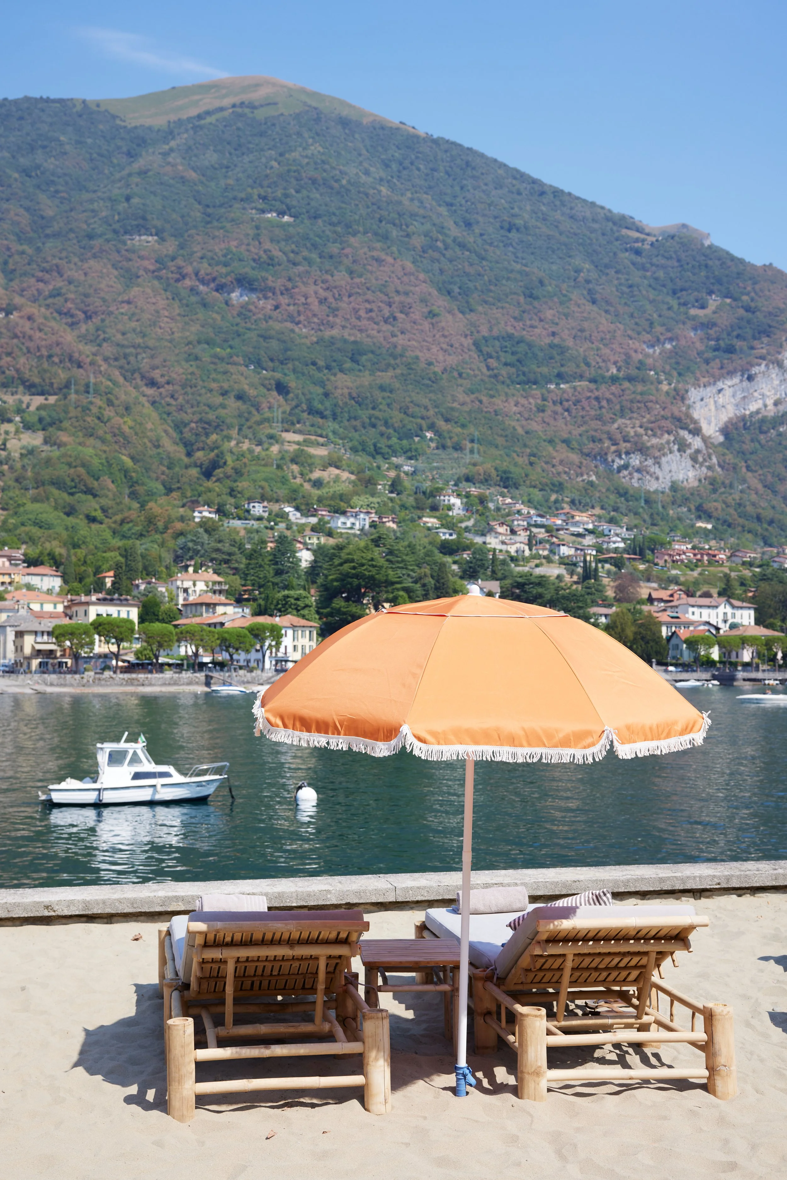 Beach scene with two wooden lounge chairs and an orange parasol on the sand, with a water body, boats, and a mountain in the background.