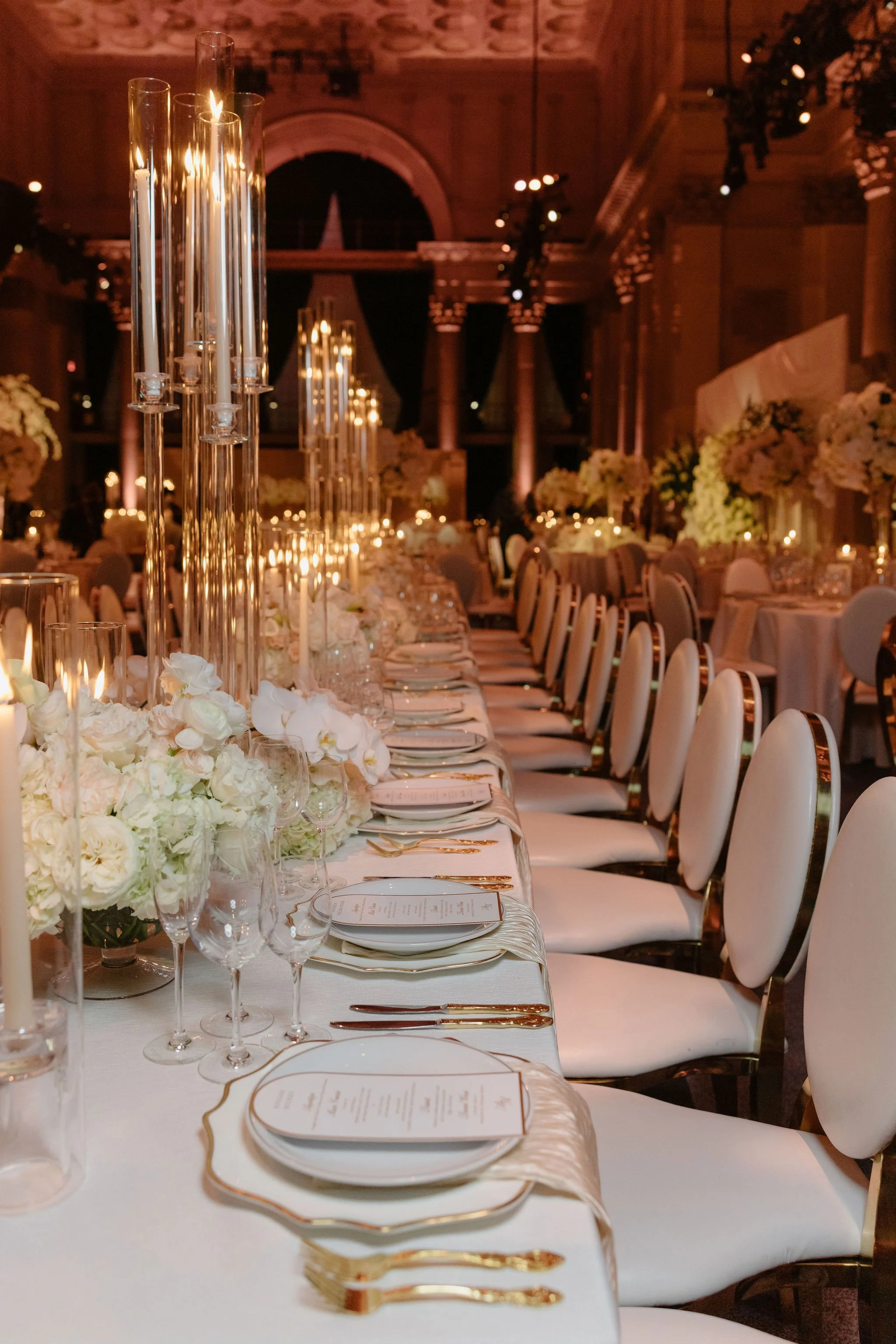 Elegant banquet hall decorated with tall glass candle holders, white floral centerpieces, and set tables with white linens, gold utensils, and glassware, preparing for a formal event or wedding reception.
