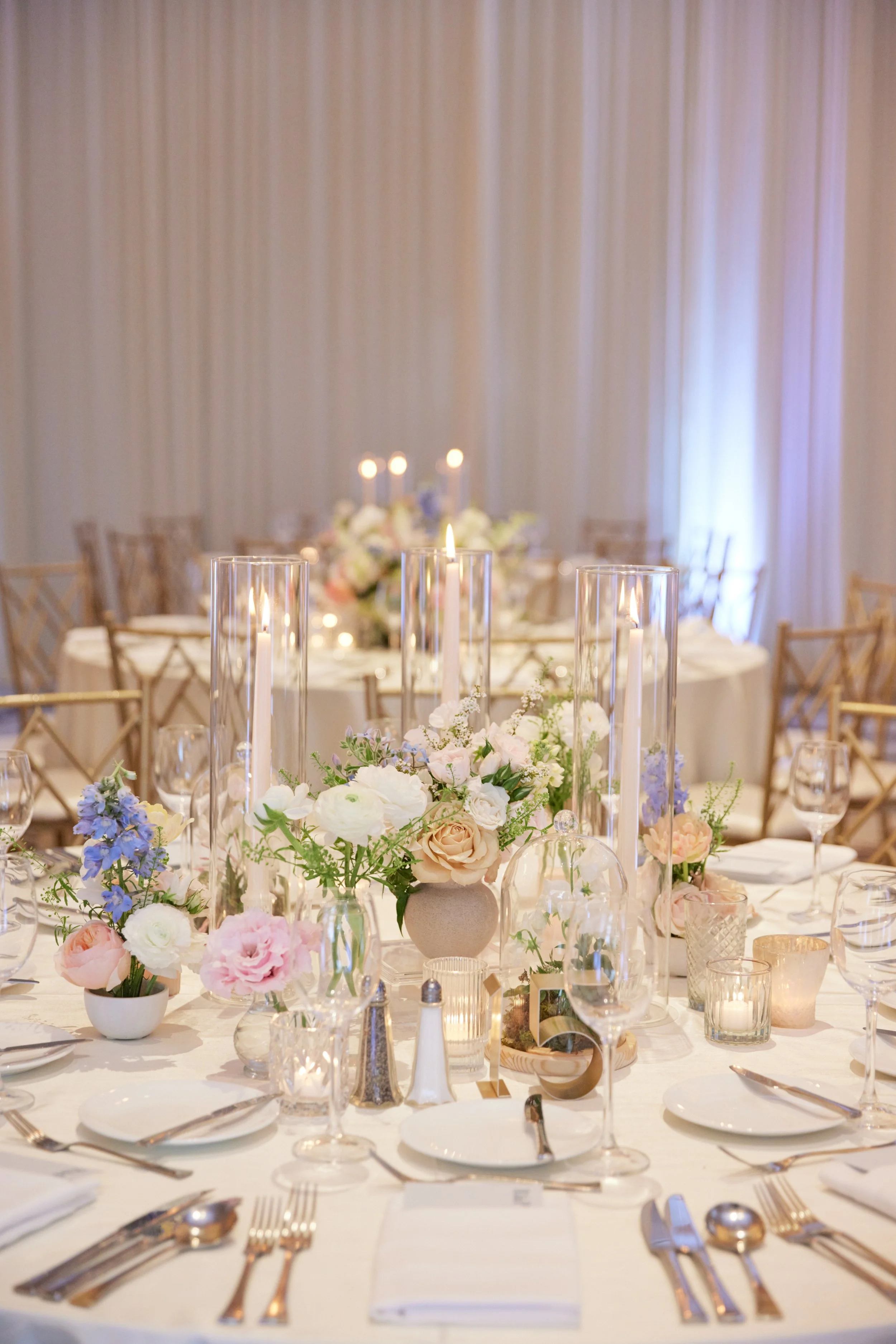 Elegant banquet table decorated with pastel-colored flowers, tall candles in glass holders, and neatly arranged tableware, set for a formal event in a decorated hall.