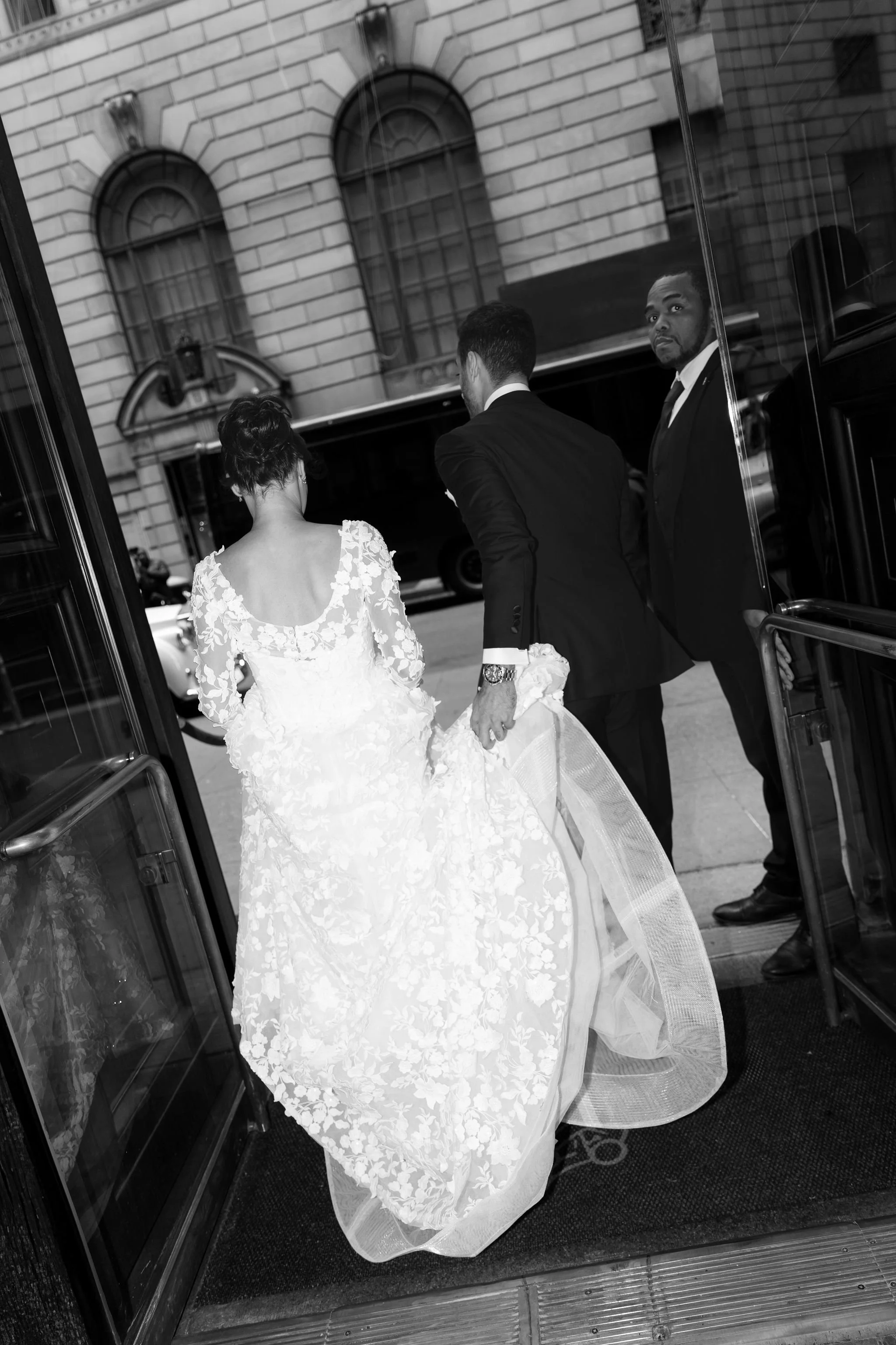 A bride in a white wedding gown with floral lace and a full skirt, holding the hand of a groom in a dark suit, as they exit a building. A man in a dark suit stands nearby, looking at the camera. The scene is reflected in a glass doorway revealing a c
