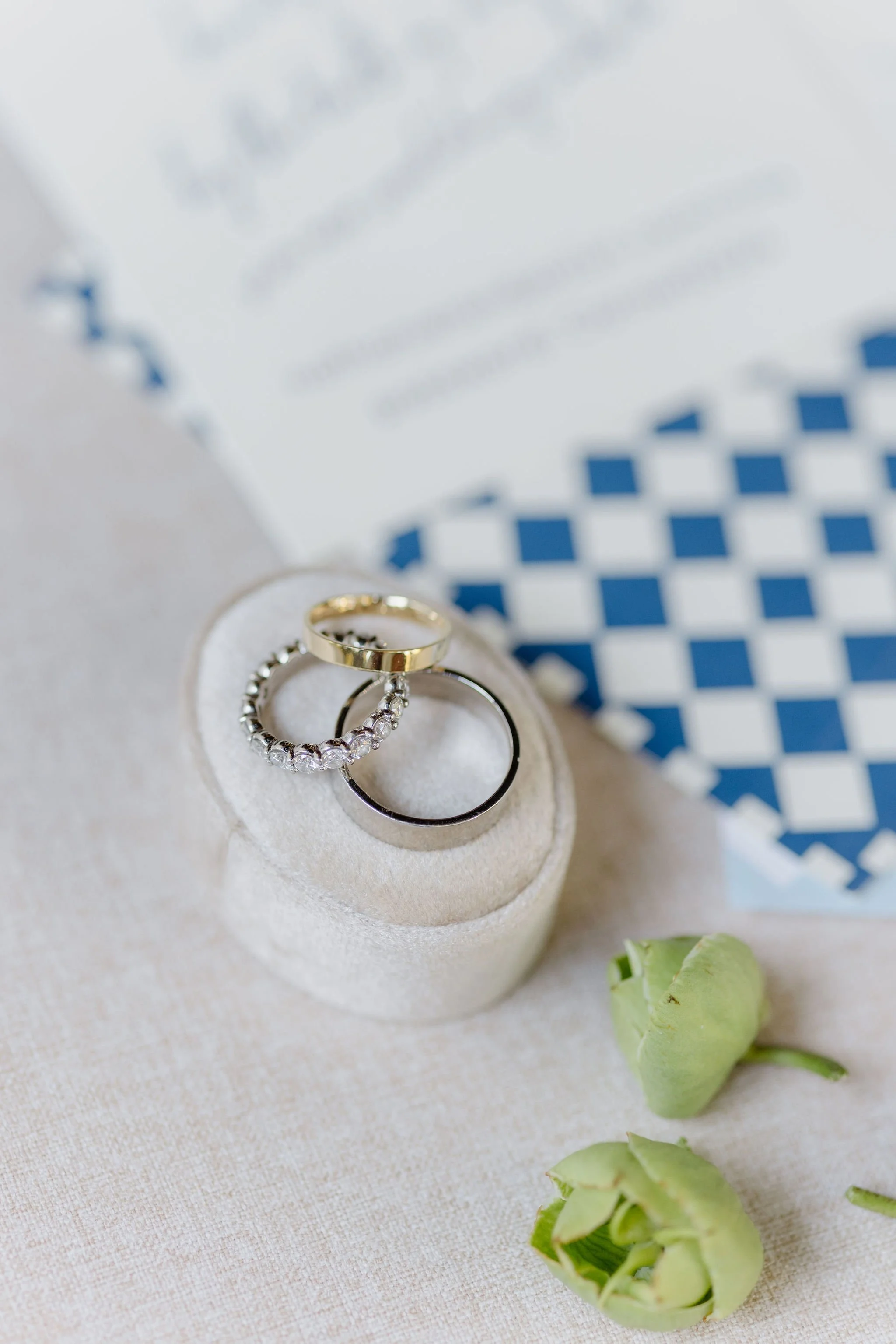 Close-up of three wedding rings, two with gems, placed on a white ring holder. Green rosebuds are visible in the bottom right corner, and a patterned blue and white paper is in the background.