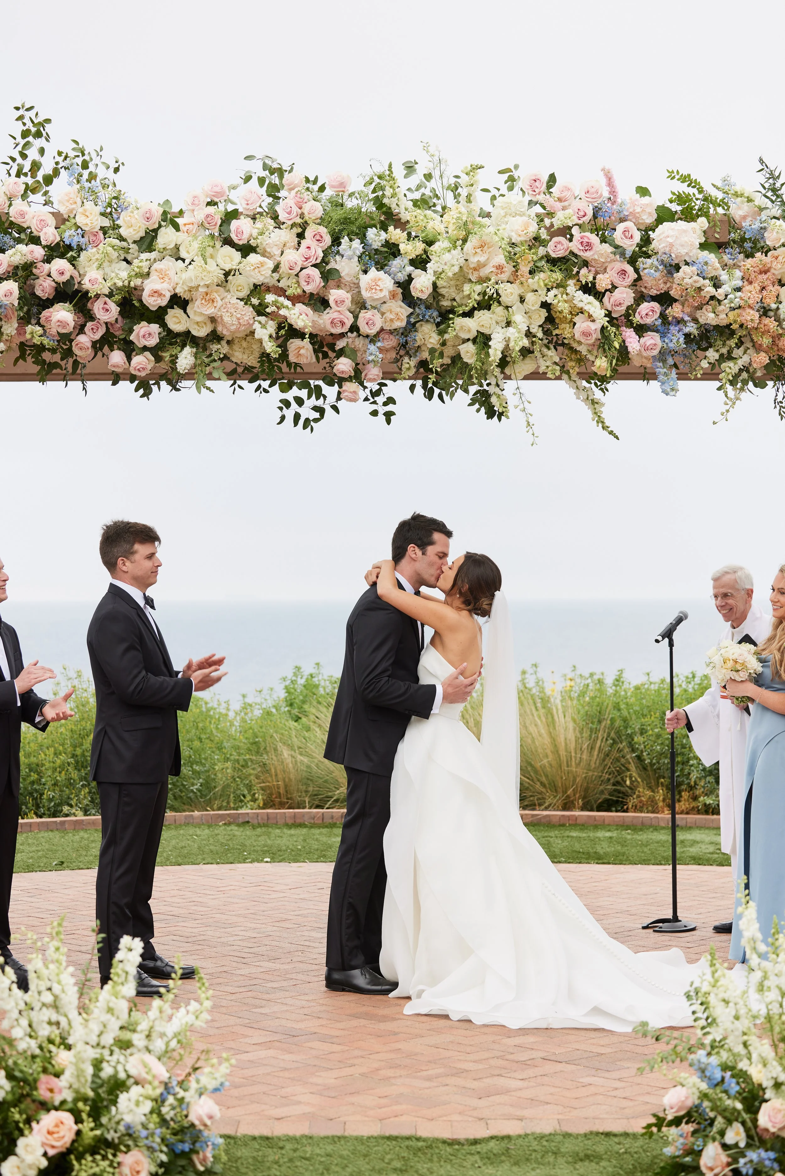 A wedding ceremony outdoors with a couple kissing under a floral arch, surrounded by family and friends clapping. The setting includes lush greenery and flowers with the ocean visible in the background.