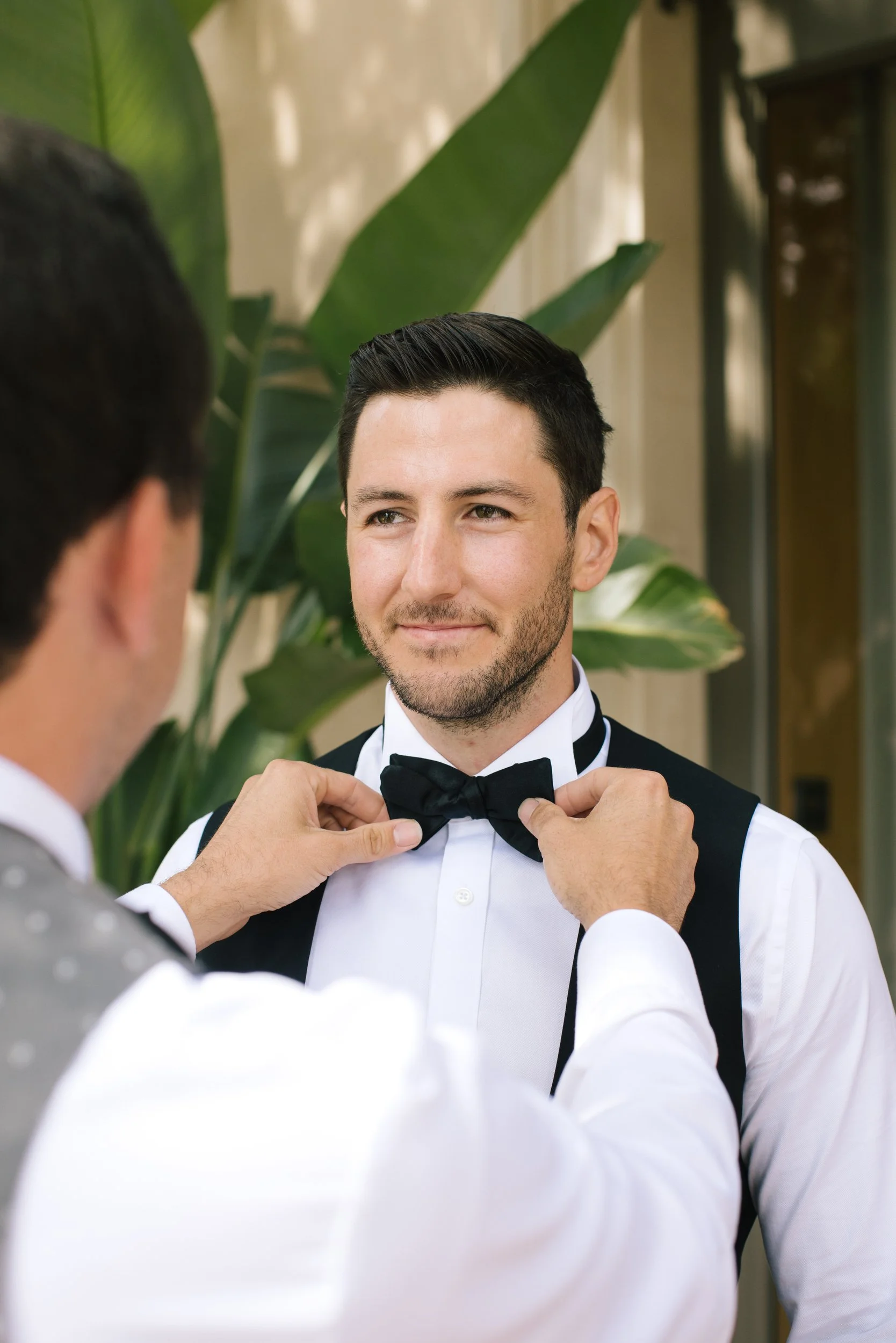 A man dressed formally in a tuxedo is being helped with his black bow tie by another man in front of large green plants.