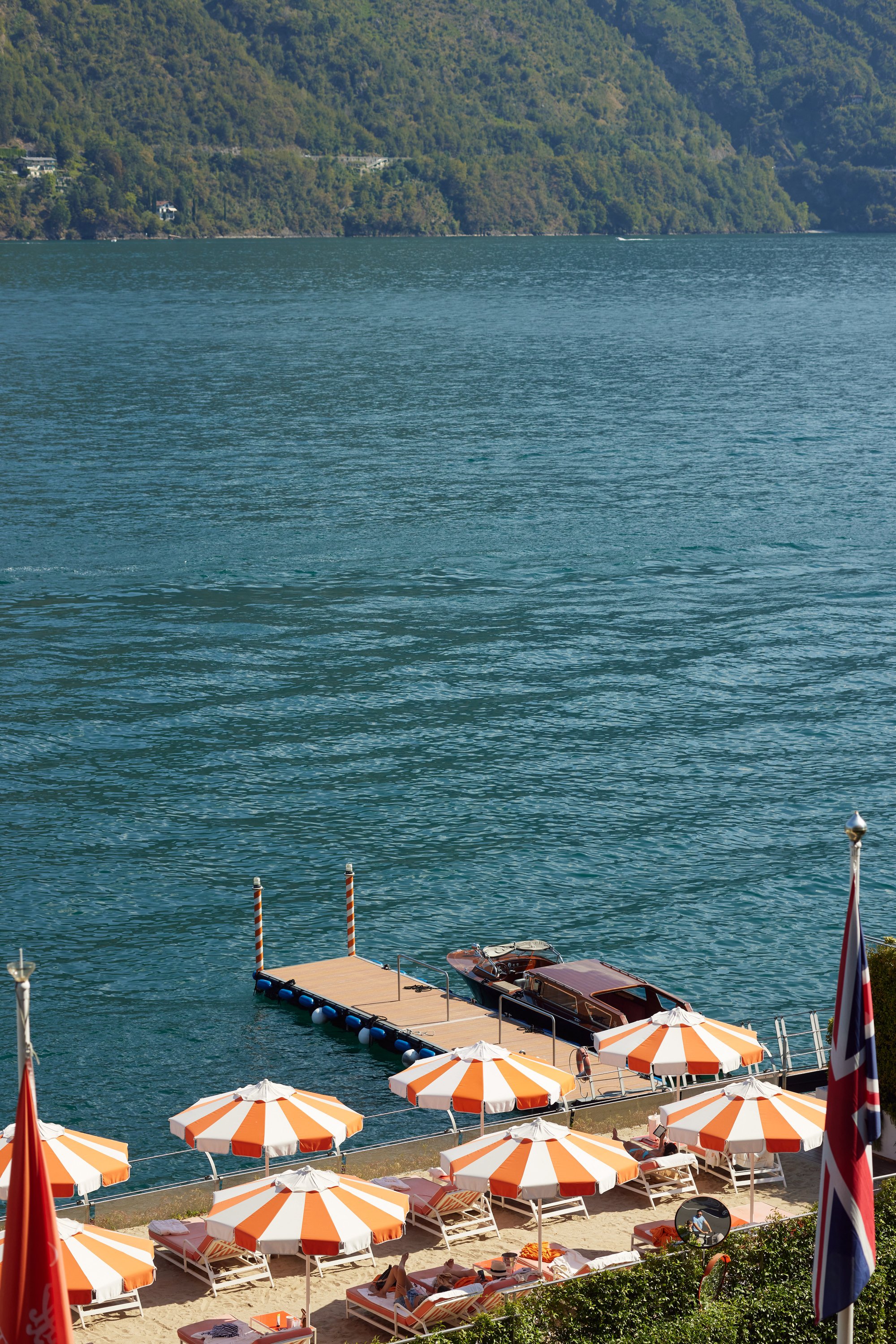 Dock with boats near orange and white striped umbrellas on a beach, overlooking a lake with green forested mountains in the background.