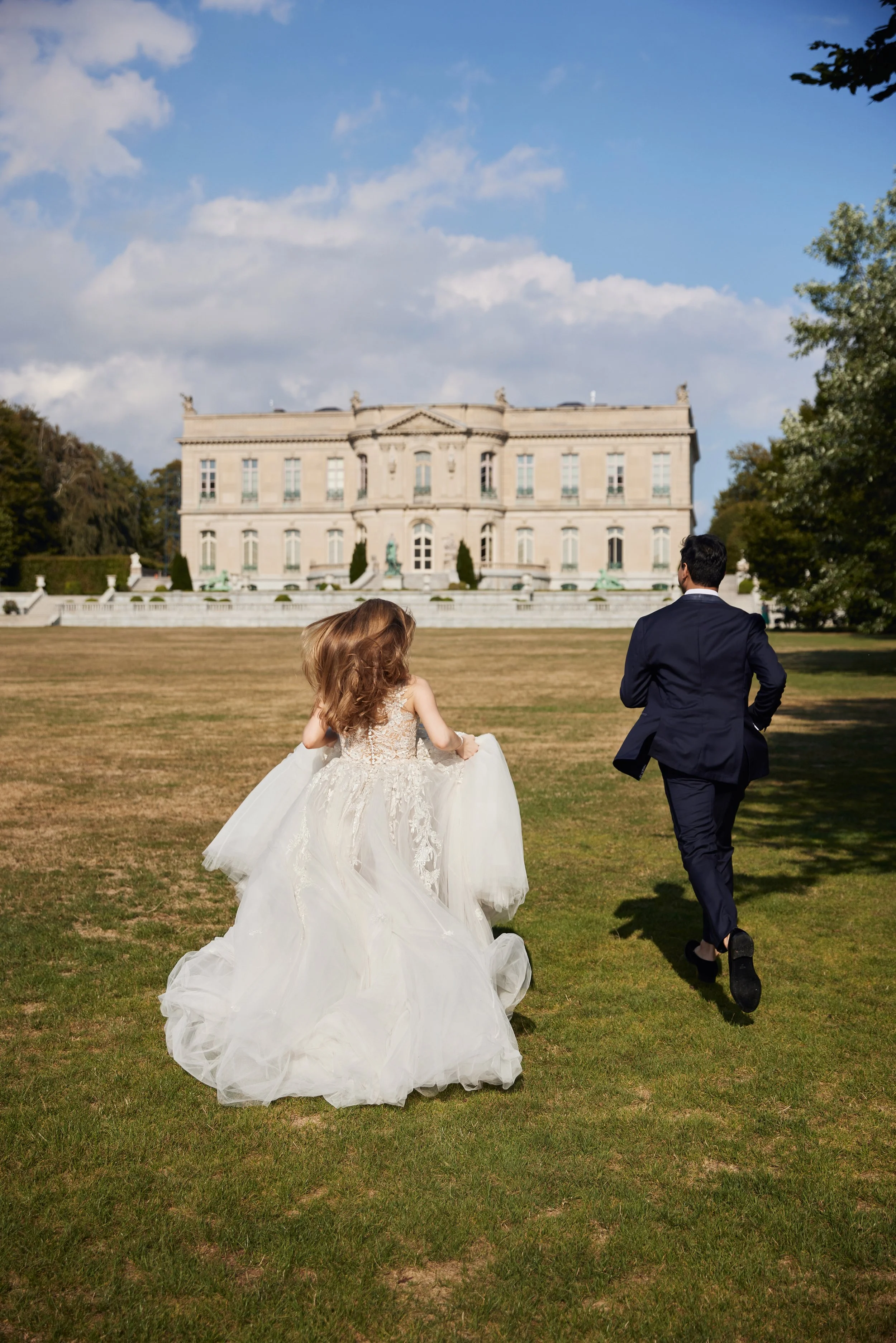 A bride and groom running towards a large historic building or mansion, on a grassy lawn with trees and a partly cloudy sky in the background.