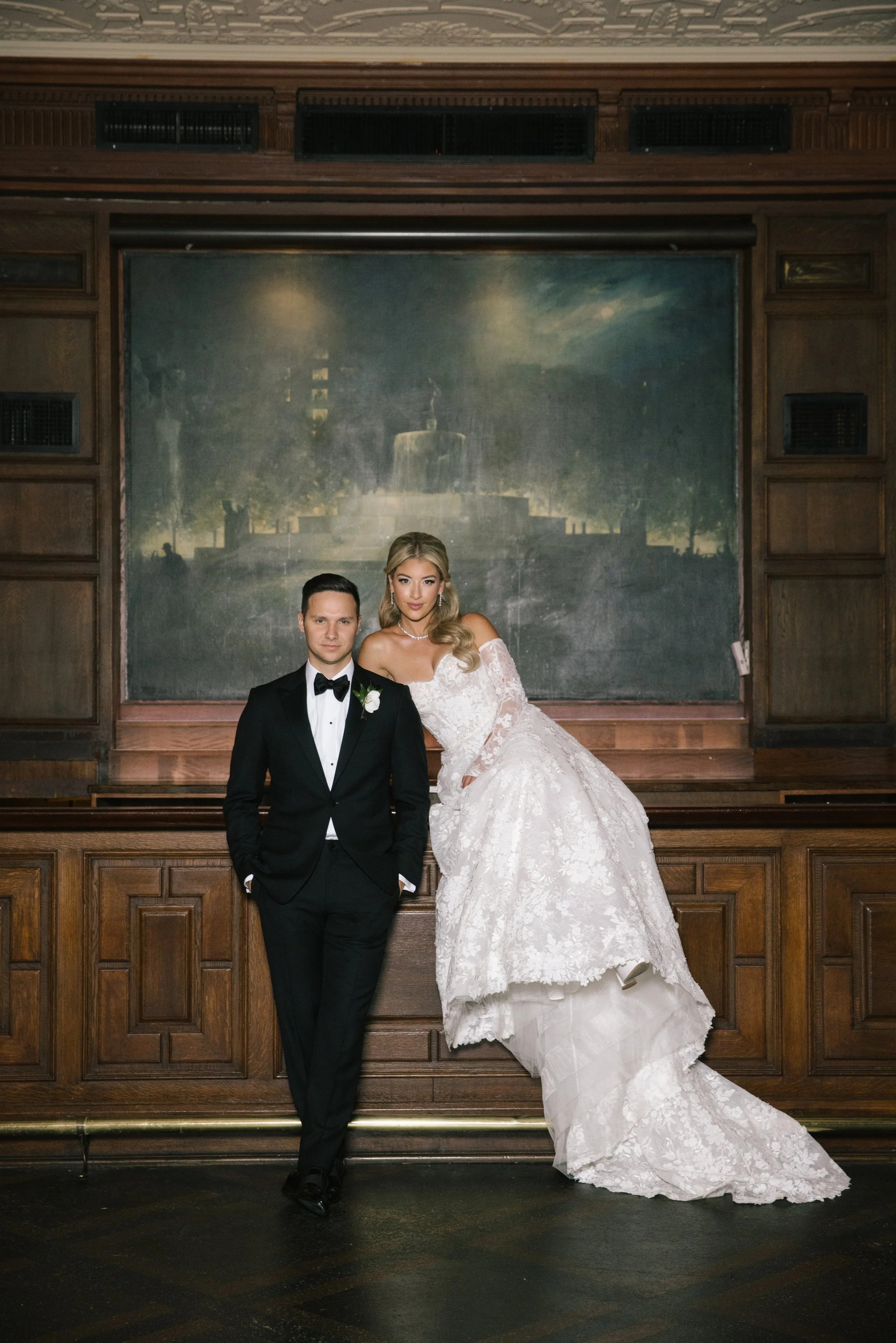 A bride and groom stand together in front of a wooden backdrop in a formal setting. The groom is dressed in a black tuxedo with a white shirt and black bow tie, with his hands in his pockets. The bride is wearing a white lace wedding gown with an off-the-shoulder neckline, and has long, wavy blonde hair. She is standing slightly behind the groom, leaning towards him.