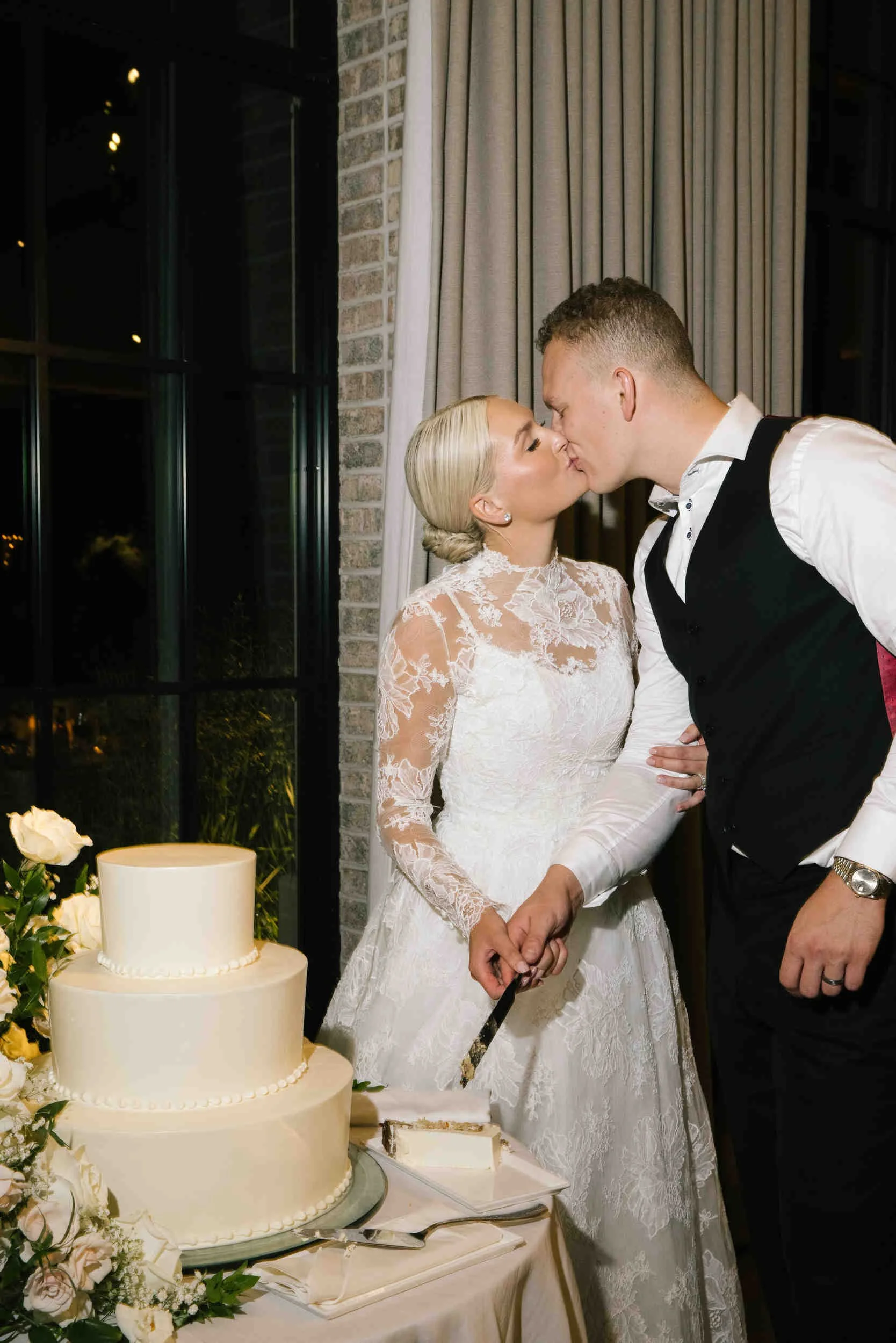 A bride and groom kissing during their wedding reception. The bride is wearing a white lace wedding gown, and the groom is in a black vest and white shirt. They are holding hands over a table with a three-tier wedding cake decorated with white flower