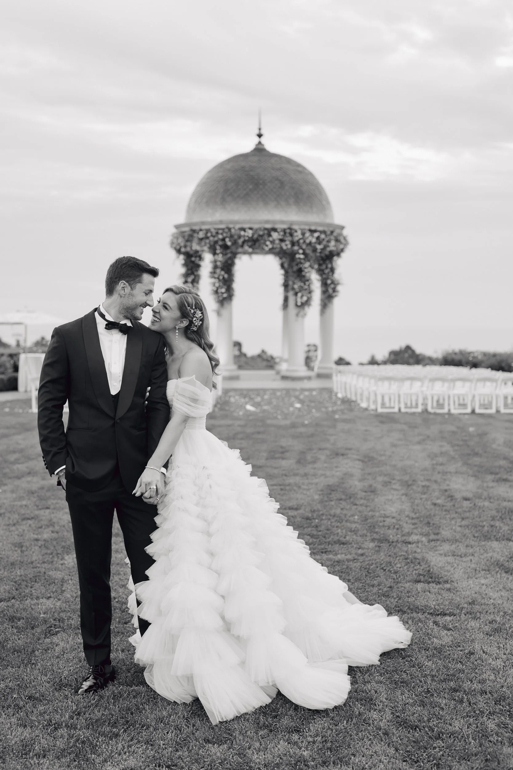 A wedding couple, dressed in formal attire, smiling and holding hands outdoors, with an ornate gazebo with a domed roof in the background.