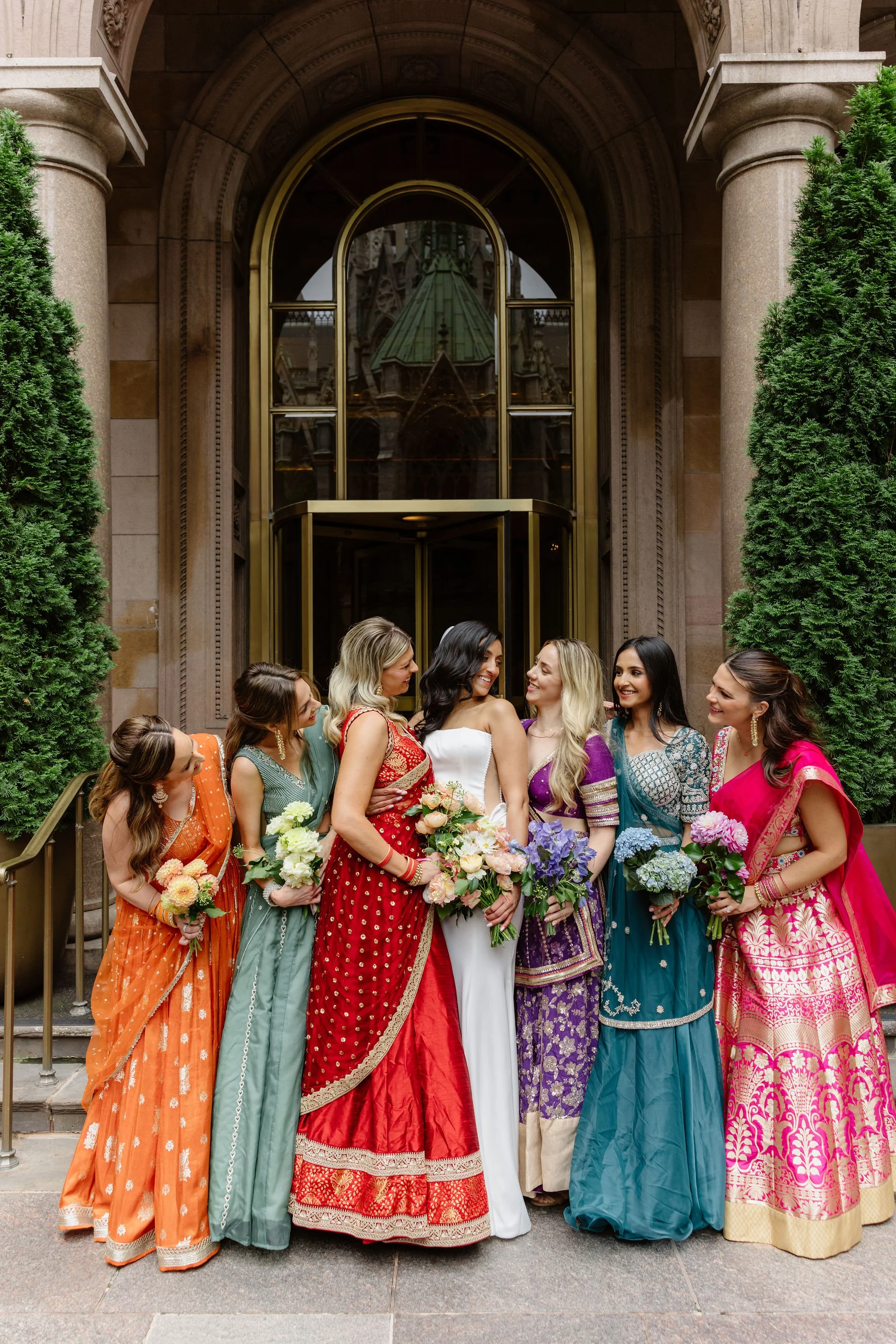 Group of women in colorful traditional Indian dresses holding flowers, standing on steps in front of a building with a large arched entrance and greenery on either side.