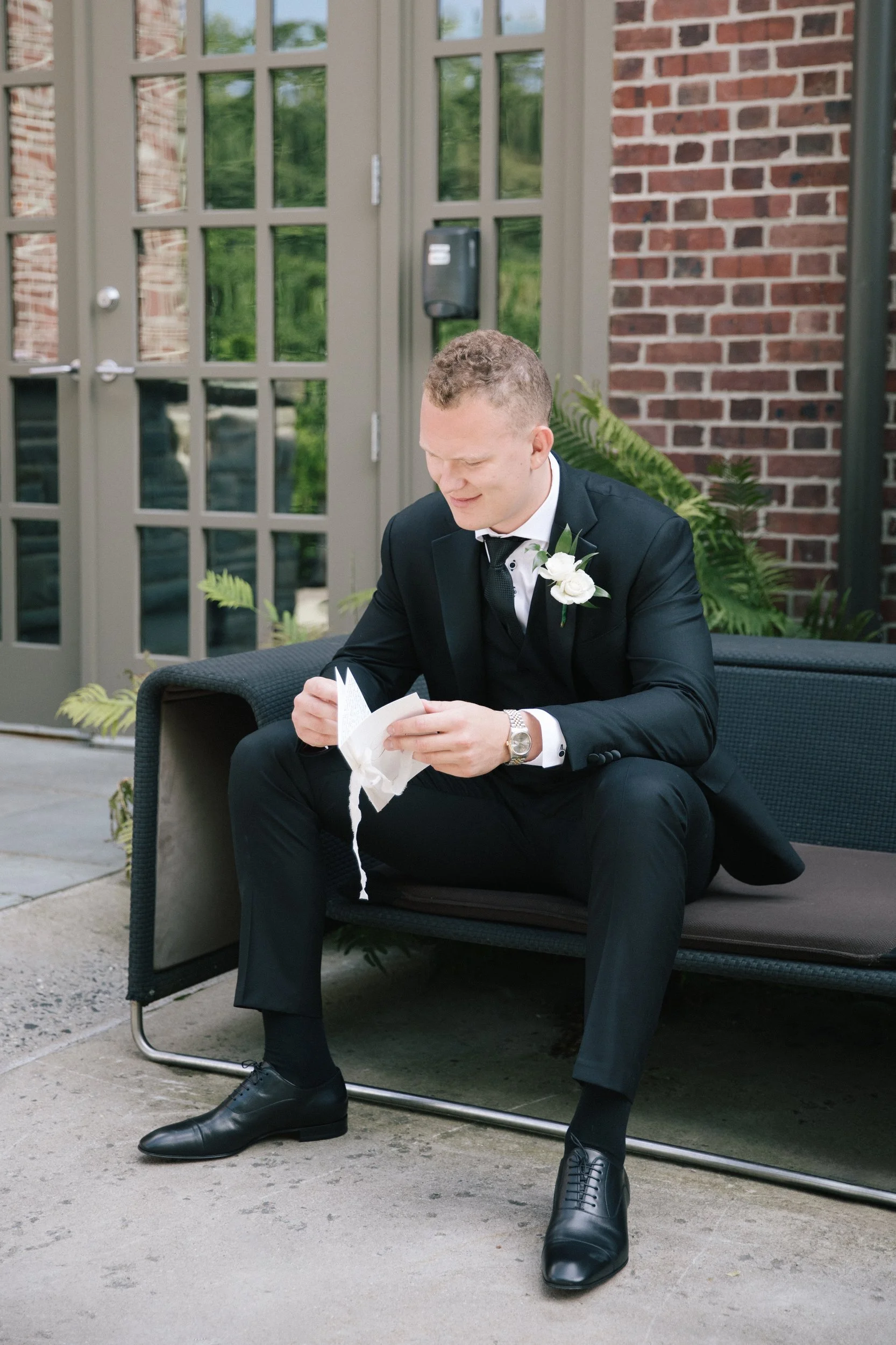 A young man dressed in a black tuxedo with a white shirt, black tie, and boutonniere, sitting on an outdoor lounge chair, looking at a white paper, with a brick building and greenery behind him.