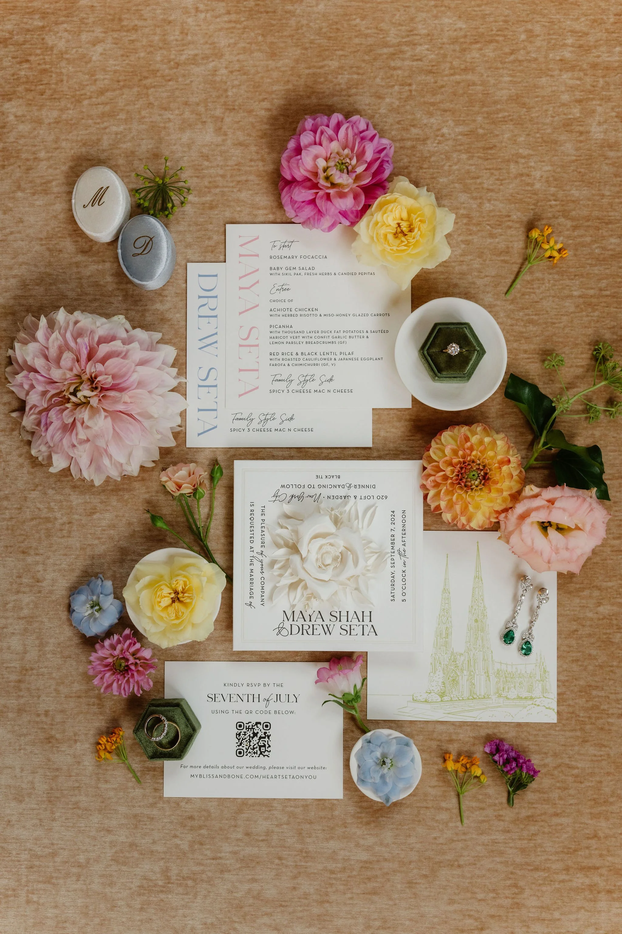Flat lay of wedding invitation suite decorated with pink, yellow, and orange flowers, green leaves, and earrings with green gemstones, on a wooden surface.