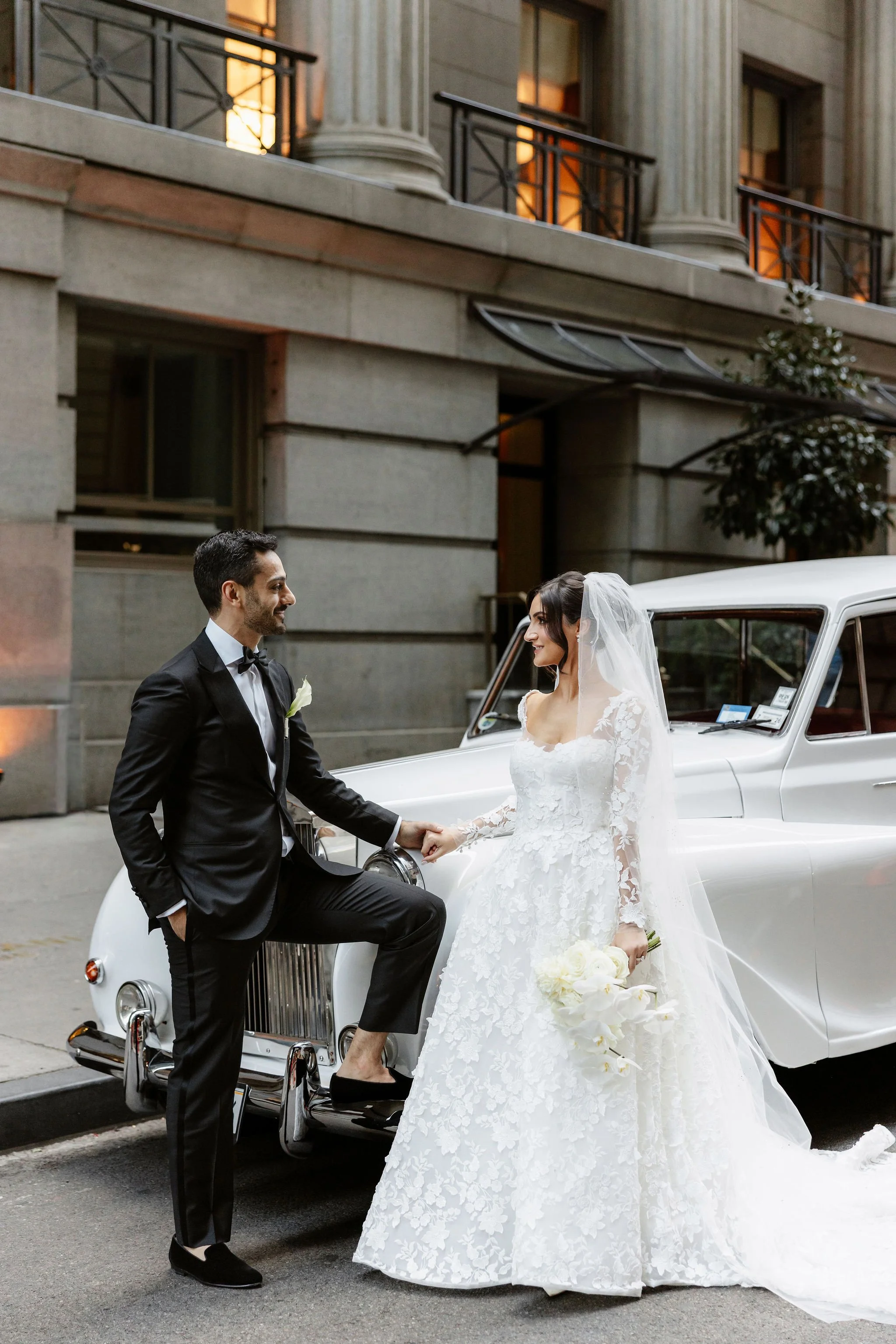 A bride and groom in wedding attire exchange vows outside a building, with the groom leaning on a vintage white car, holding the bride's hand. The bride is holding a bouquet of white flowers.
