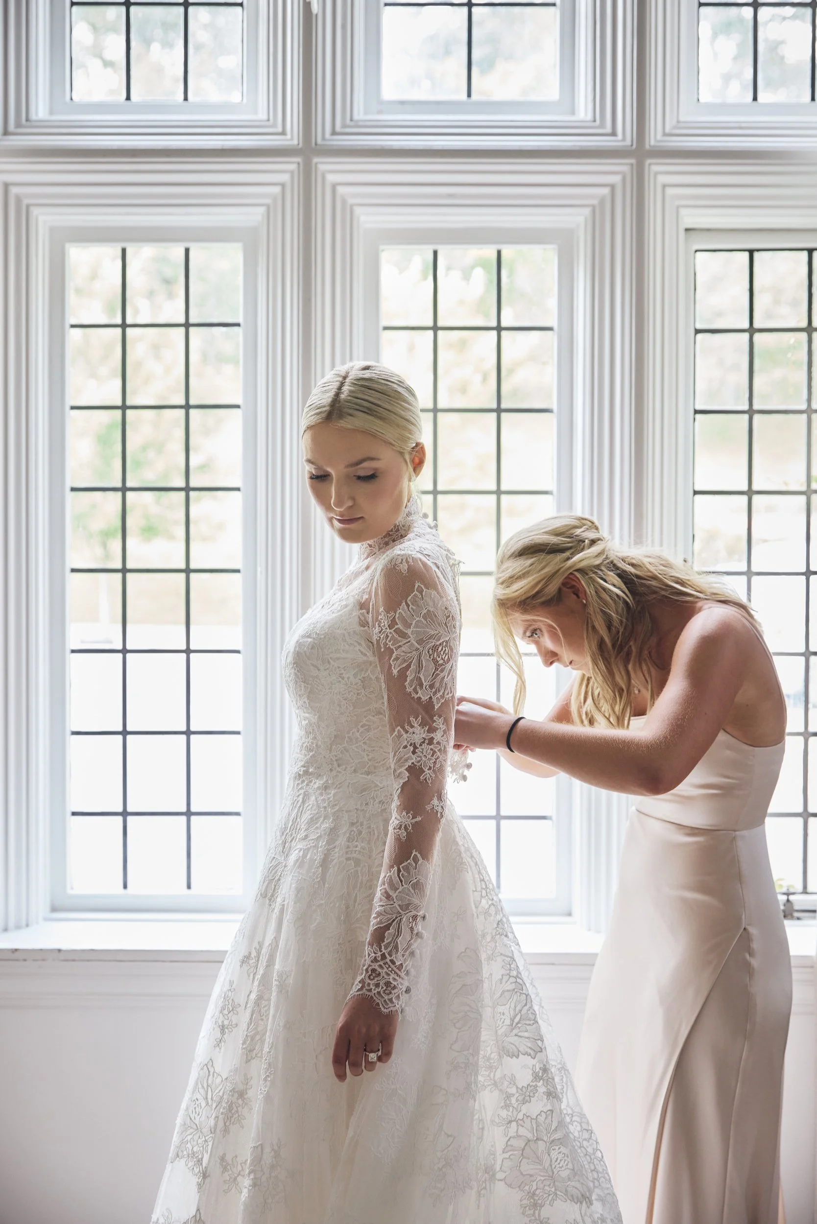 A bride in a white lace wedding dress with long sleeves is standing in front of a large window, while a woman in a strapless dress is assisting her, possibly adjusting her gown or jewelry.