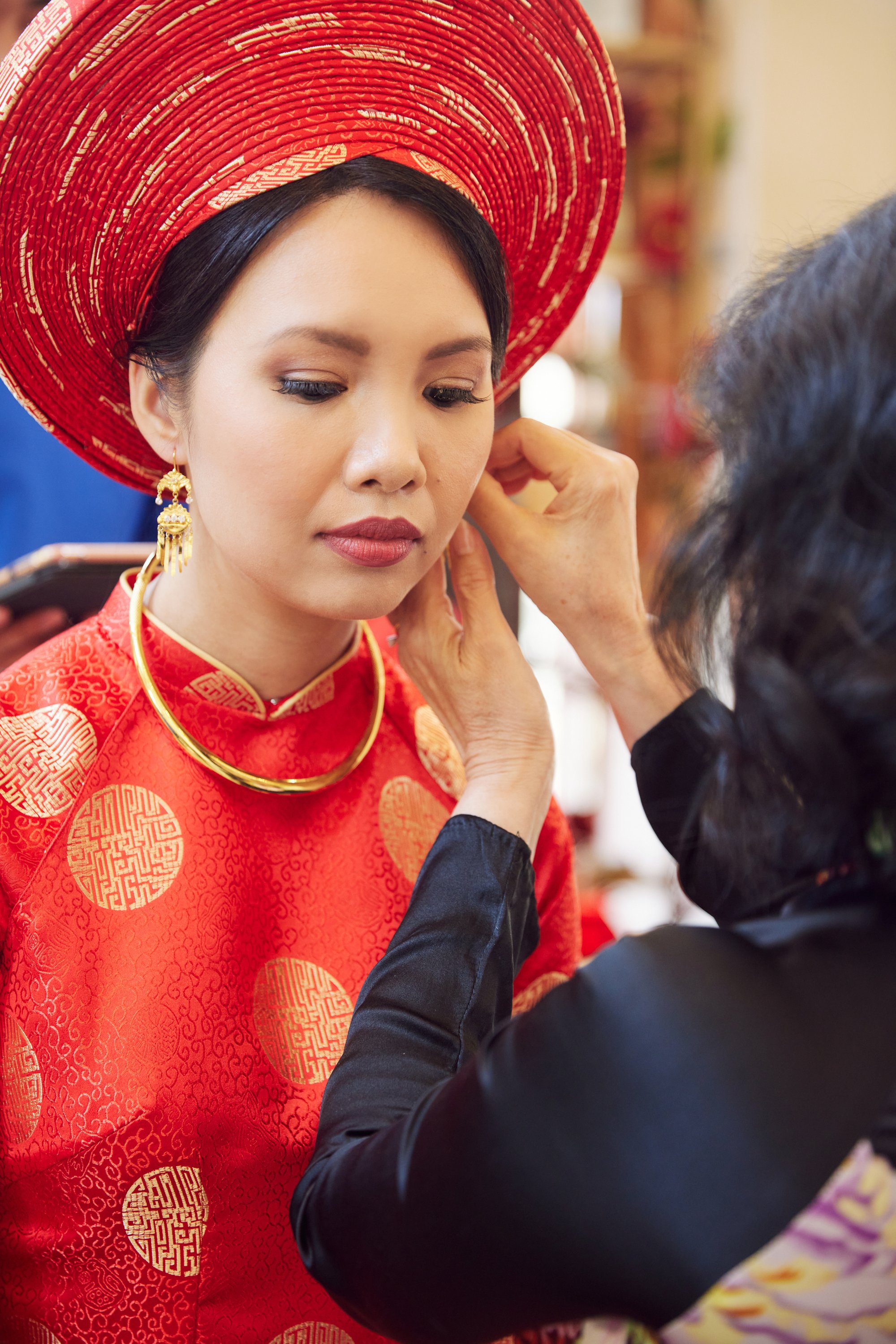 A woman dressed in traditional red Chinese wedding attire with gold patterns and a large red hat, having her makeup adjusted by another person.