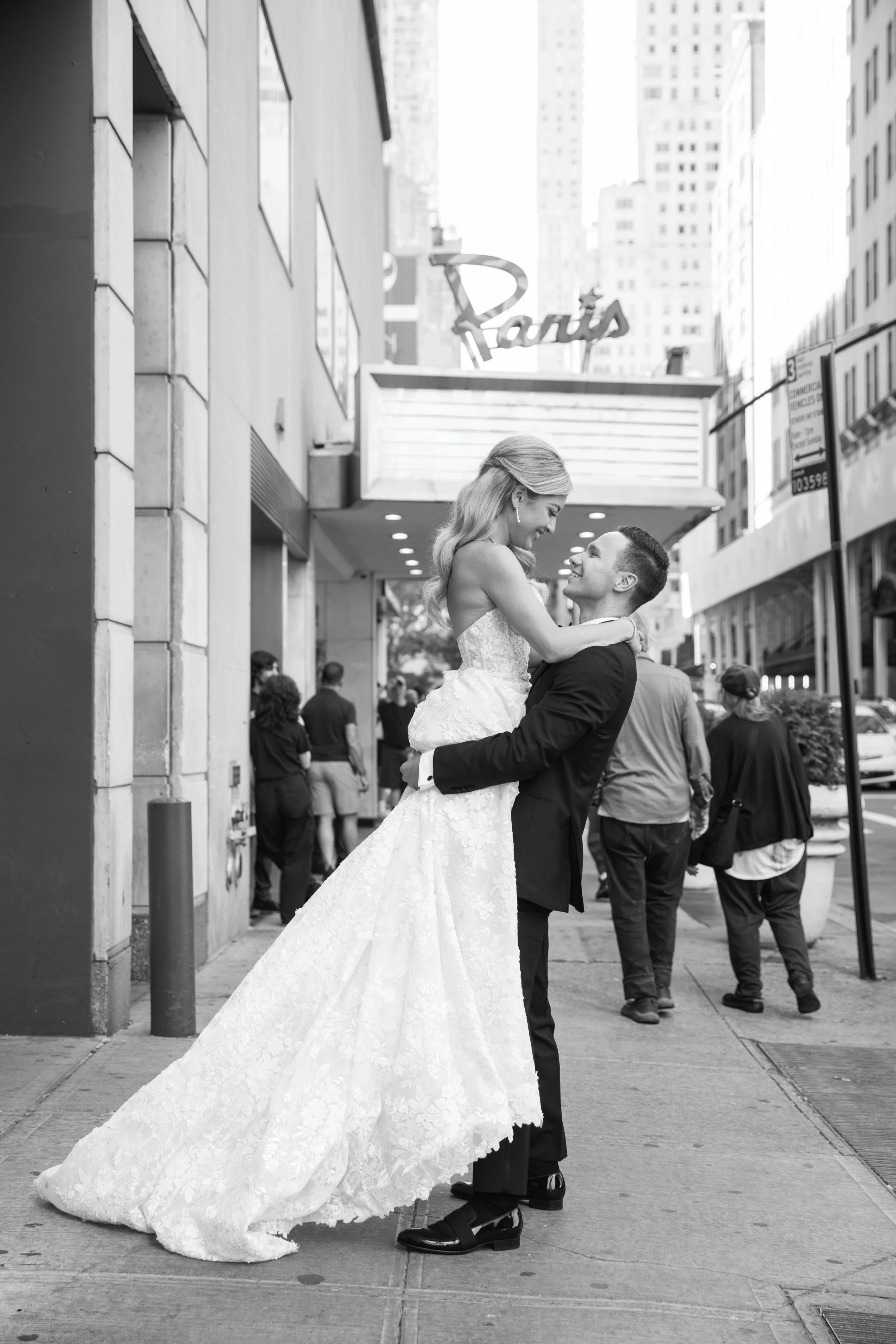 A black and white photo of a bride and groom on a city street, with the groom lifting the bride. She is wearing a strapless lace wedding gown, and he is in a dark suit. People are walking in the background and a sign for Pantages is visible above the