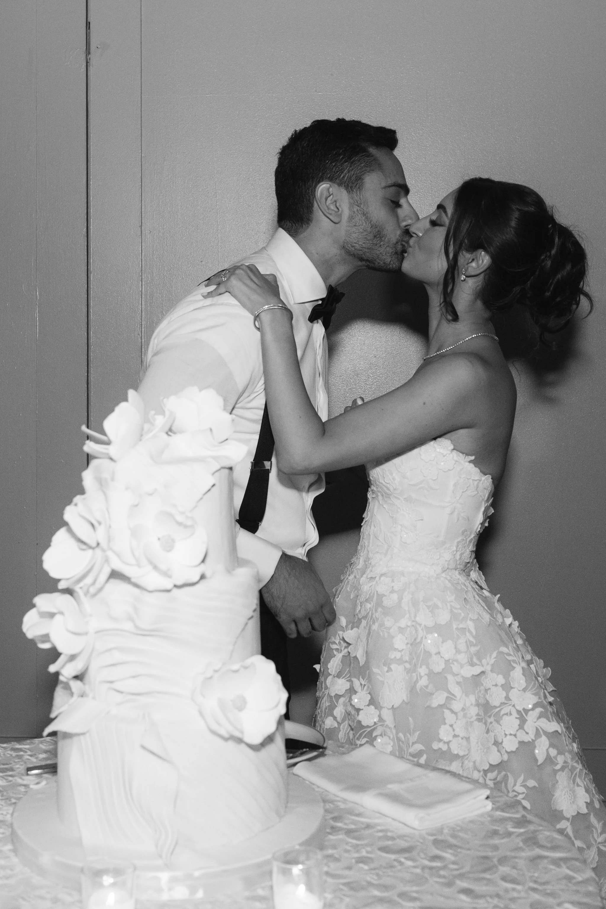 Black and white photograph of a bride and groom sharing a kiss during their wedding reception, with a multi-tiered wedding cake decorated with flowers in the foreground.