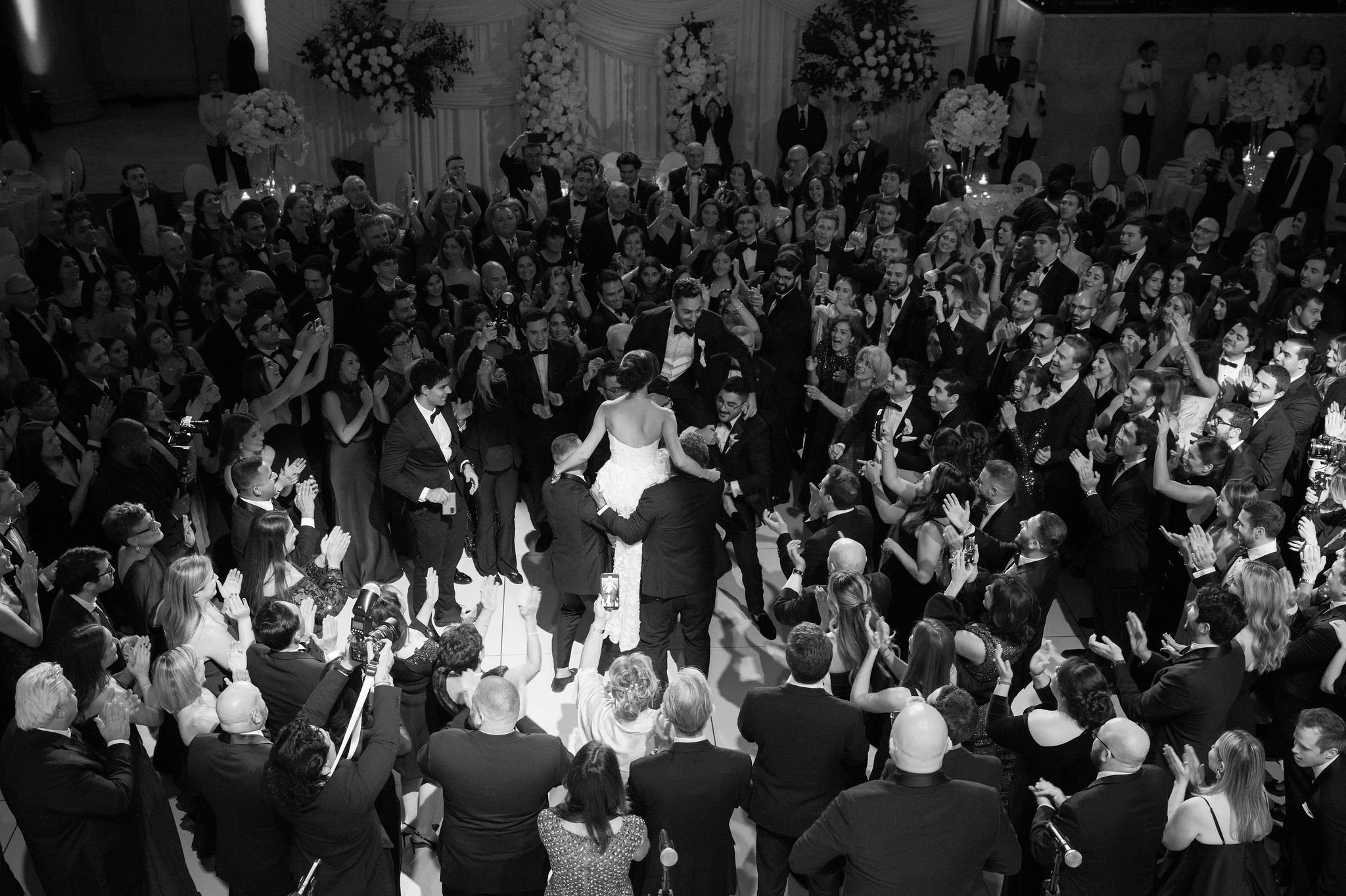 A black and white photo of a wedding reception with a large crowd of guests dancing and celebrating. The bride and groom are in the center, and there are floral arrangements and decorations in the background.