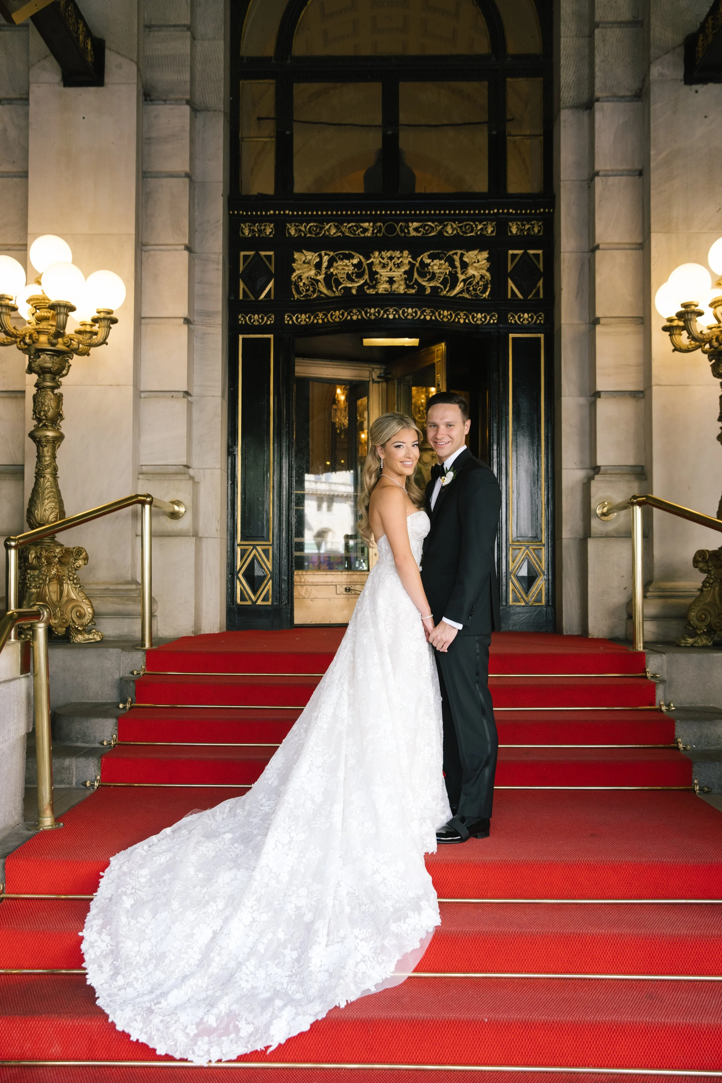 A bride and groom stand on a red carpeted staircase outside a grand building, smiling and holding hands. The bride wears a white lace wedding gown with a long train, and the groom wears a black tuxedo with a bow tie. The entrance features ornate black and gold decorative elements and brass railings, illuminated by two large, ornate brass lamps.