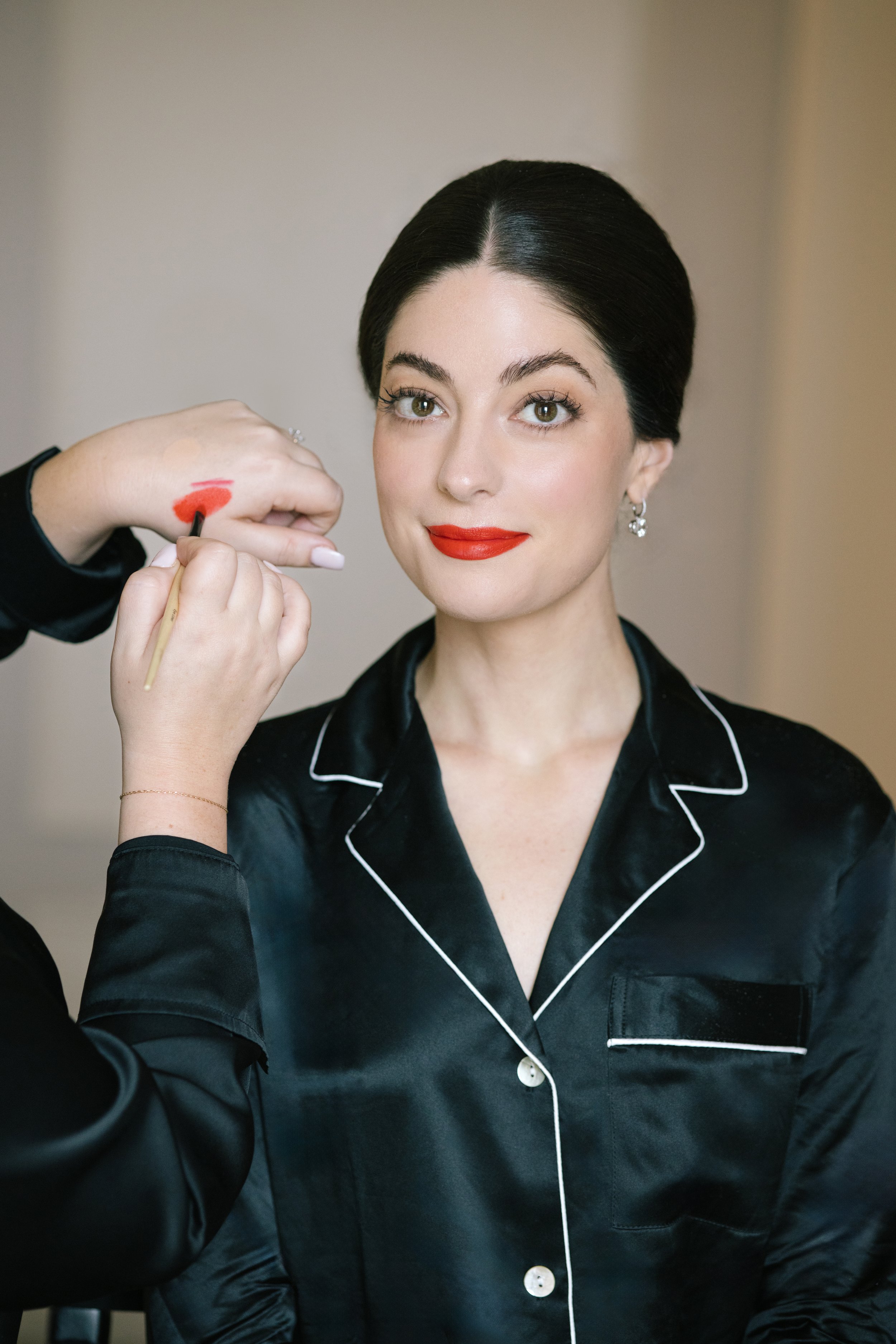 A woman with black hair and earrings having makeup applied on her cheeks, wearing a black satin silk pajama with white piping, sitting in a room with beige walls.