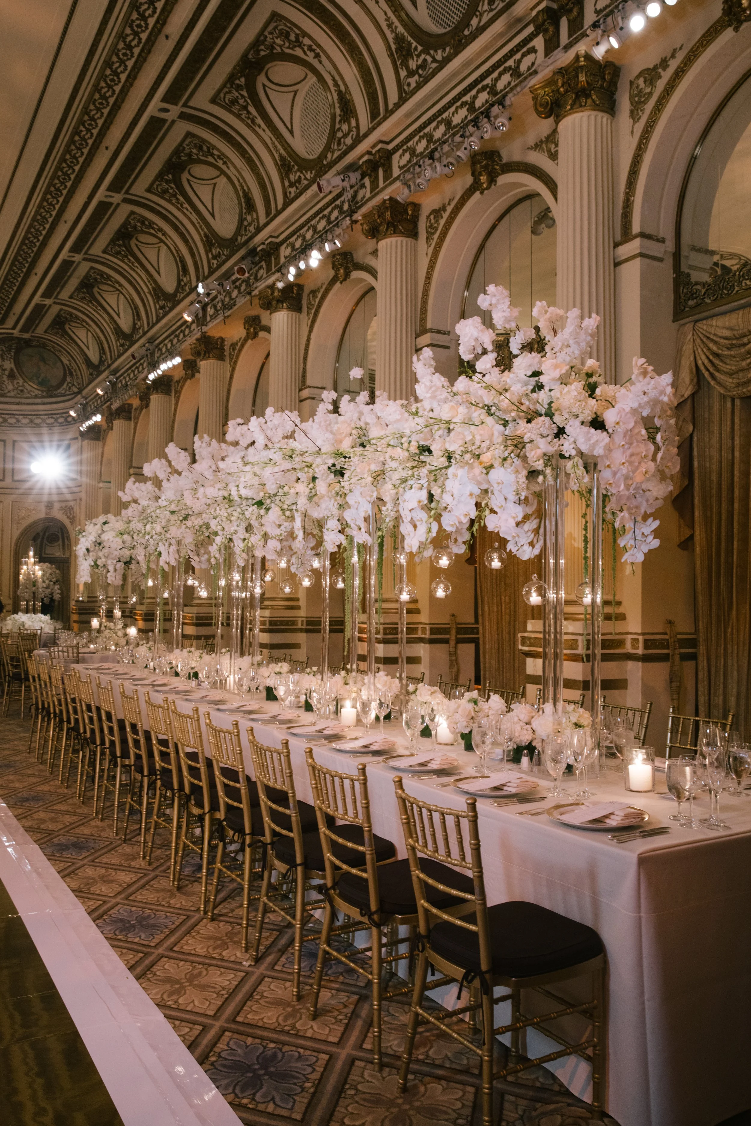 Elegant banquet hall decorated for a formal event with a long table surrounded by gold chairs, tall floral centerpieces with white and pale pink flowers, candles, and table settings, ornate ceiling with detailed moldings and columns.
