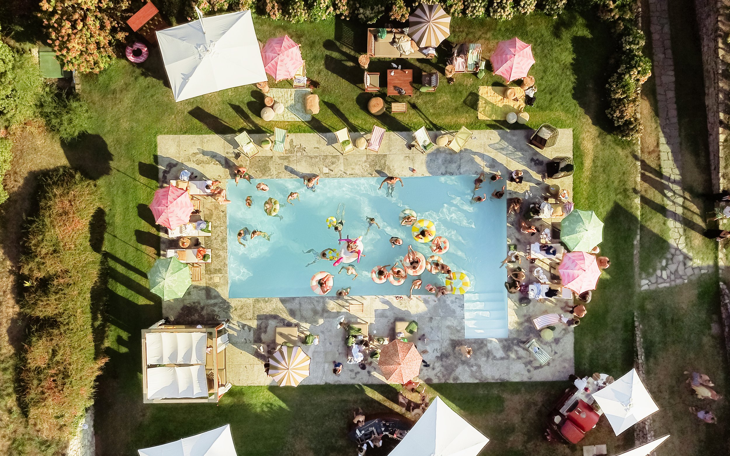 An outdoor swimming pool party with many people swimming and lounging around the pool. There are colorful umbrellas and lounge chairs, trees, and a grassy area surrounding the pool.