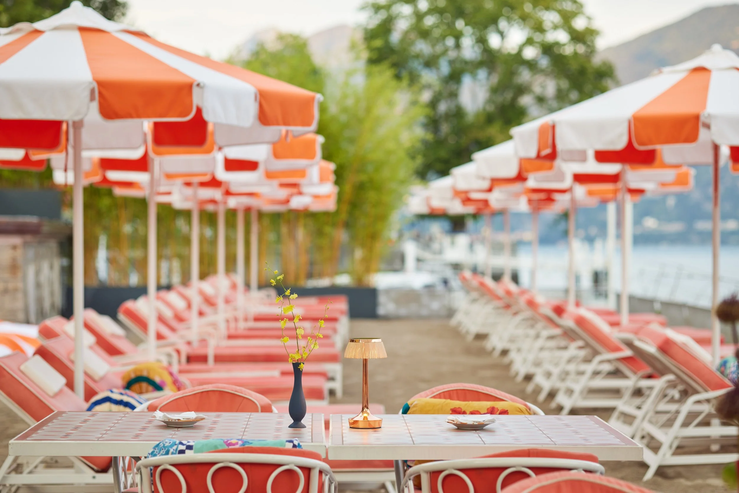 Empty outdoor patio with red and white striped umbrellas, white tables with pink checkered tablecloths, and red chairs with colorful cushions. Centered on a table is a blue vase with yellow flowers and a gold decorative lamp. The background shows gre