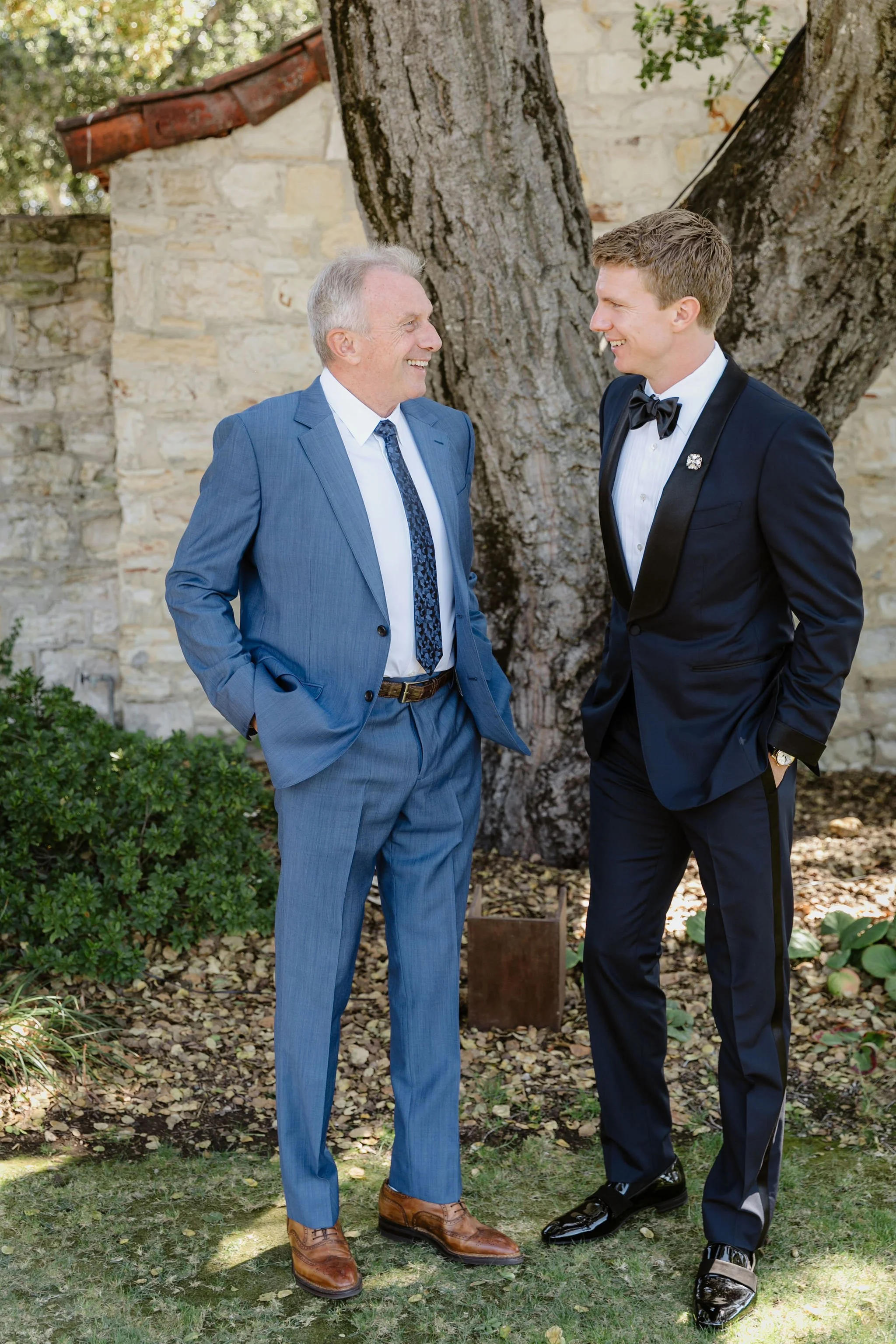 Two men in formal suits are smiling and chatting outdoors near a large tree, with a stone wall in the background.