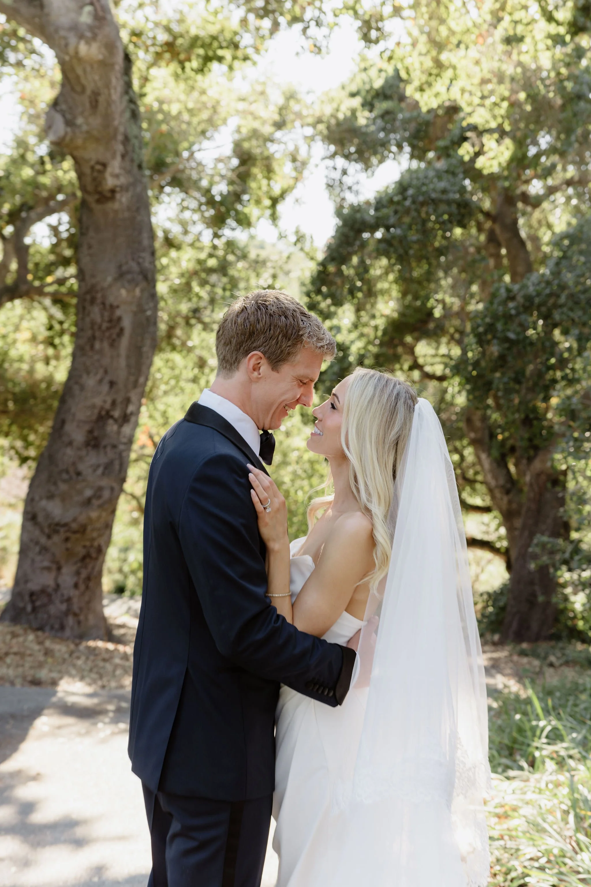 A wedding couple, a man in a black tuxedo and a woman in a white wedding dress and veil, standing close in a park with trees in the background, smiling at each other.