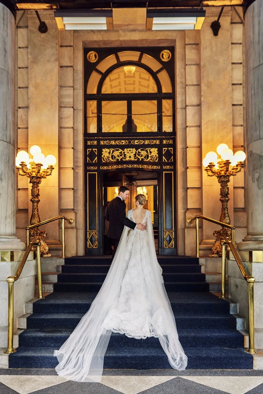 A bride in a white wedding gown and a groom in a black suit ascending the stairs at a grand, ornate building entrance, illuminated by gold and black decorative lighting fixtures.