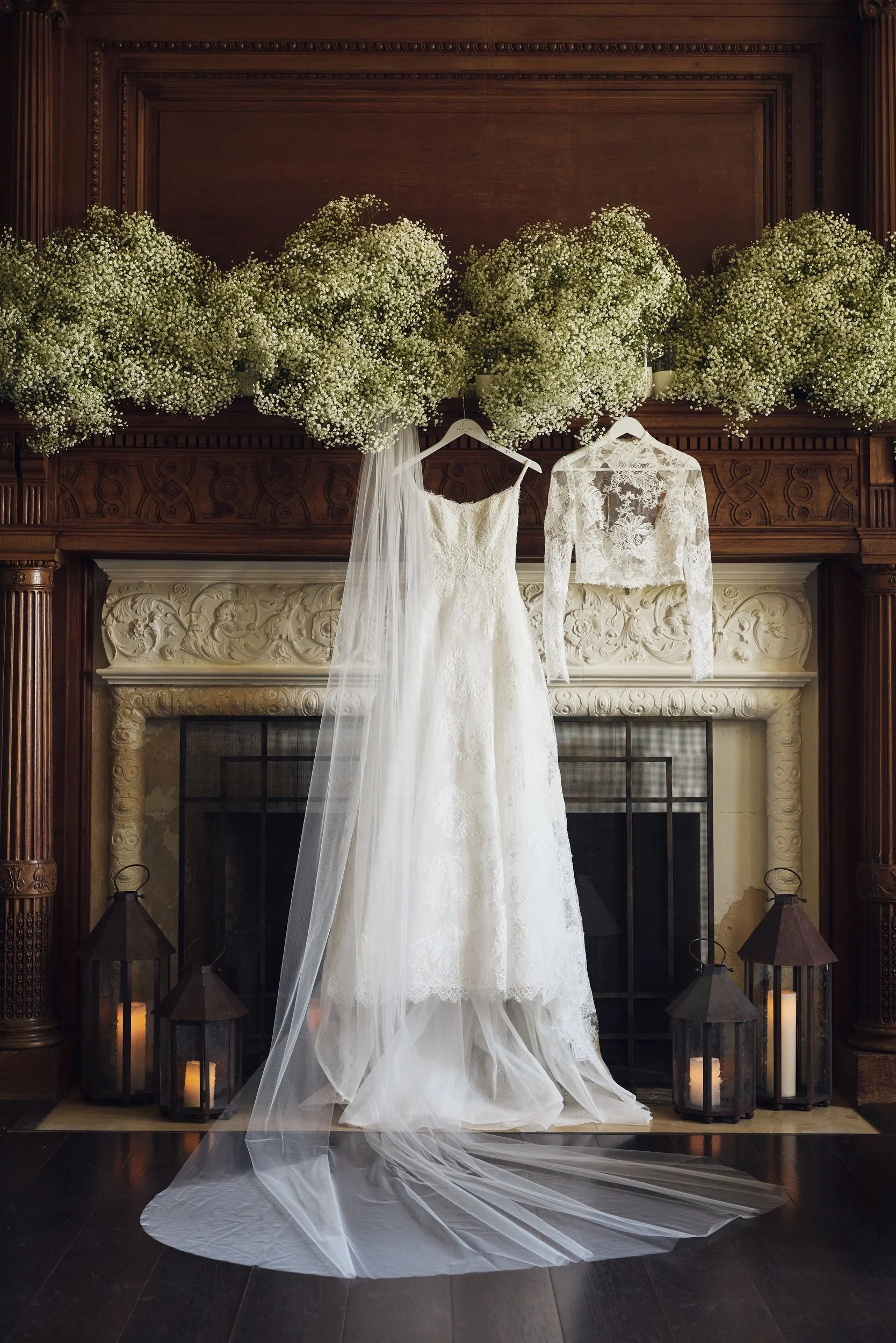 A white wedding dress hanging in front of a fireplace adorned with floral arrangements and candles.