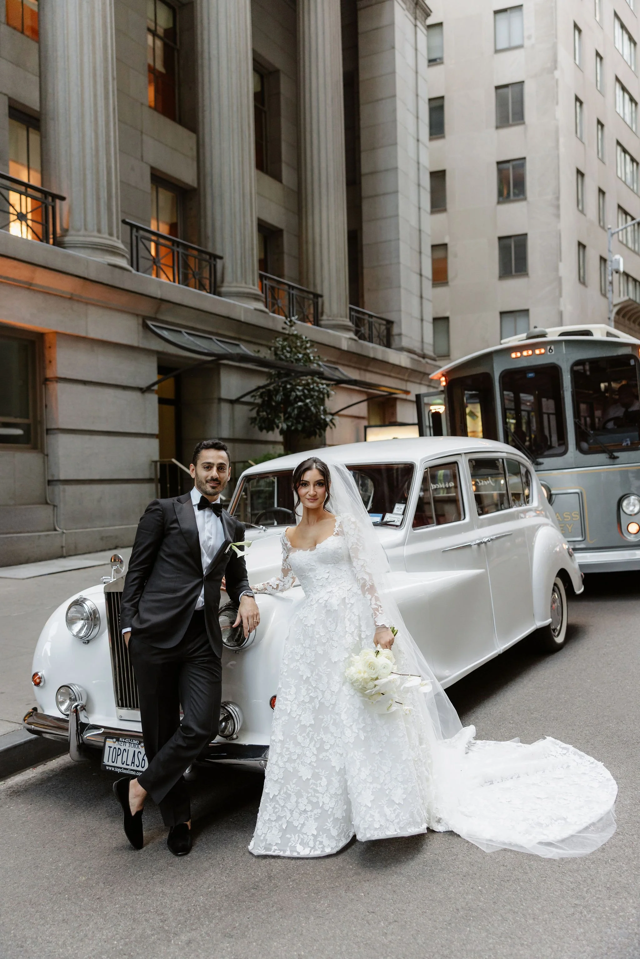 A newlywed couple in wedding attire standing beside a vintage white car on a city street. The groom is in a black tuxedo, and the bride is in a white lace wedding gown holding a bouquet of white flowers. They are smiling and posing for the photo with