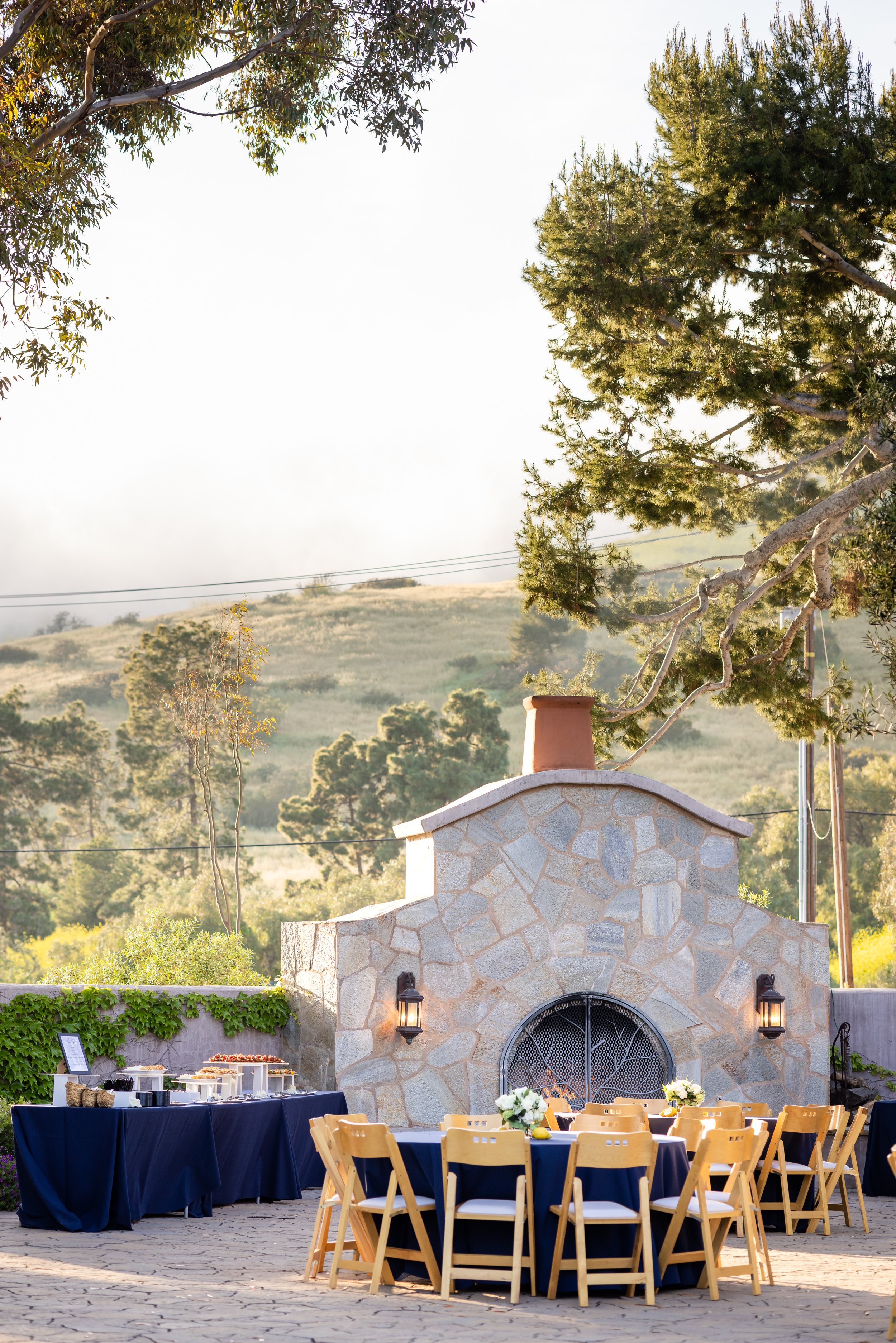 Outdoor event setup with tables covered in navy blue tablecloths, decorated with flower arrangements, in front of a stone fireplace with lanterns mounted on it, surrounded by trees and a hillside background.