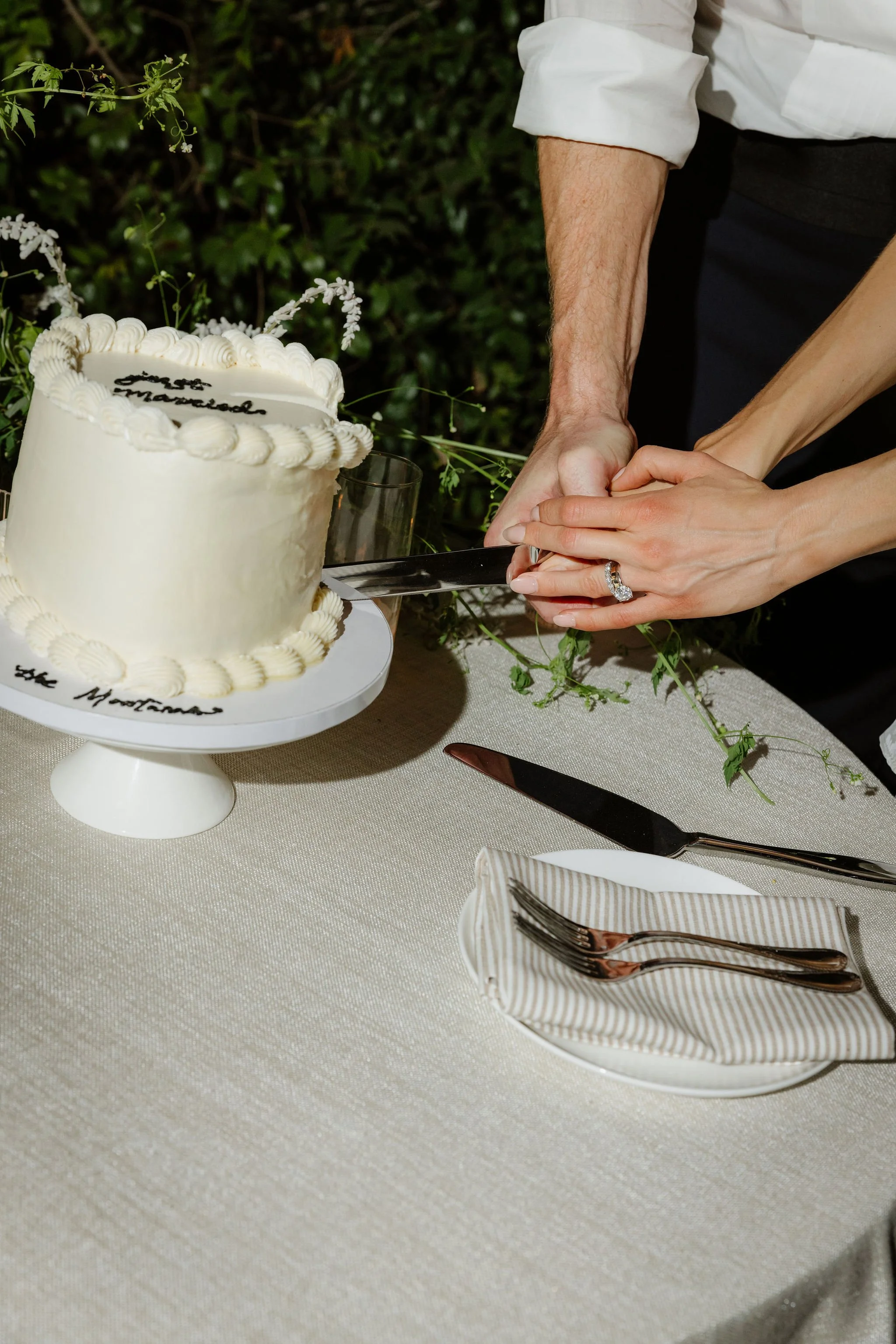A couple cutting wedding cake together at a reception. The cake is white with decorative piping and writing, set on a white cake stand on a beige tablecloth with a striped napkin and cutlery nearby.