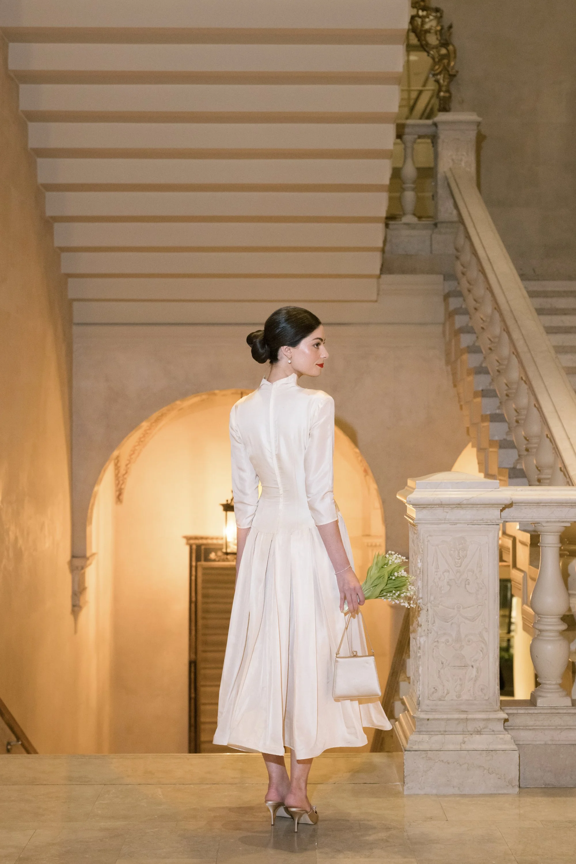A woman in a white dress holding a small bouquet of white flowers and a beige handbag, standing in an elegant interior with a staircase and arched doorway.