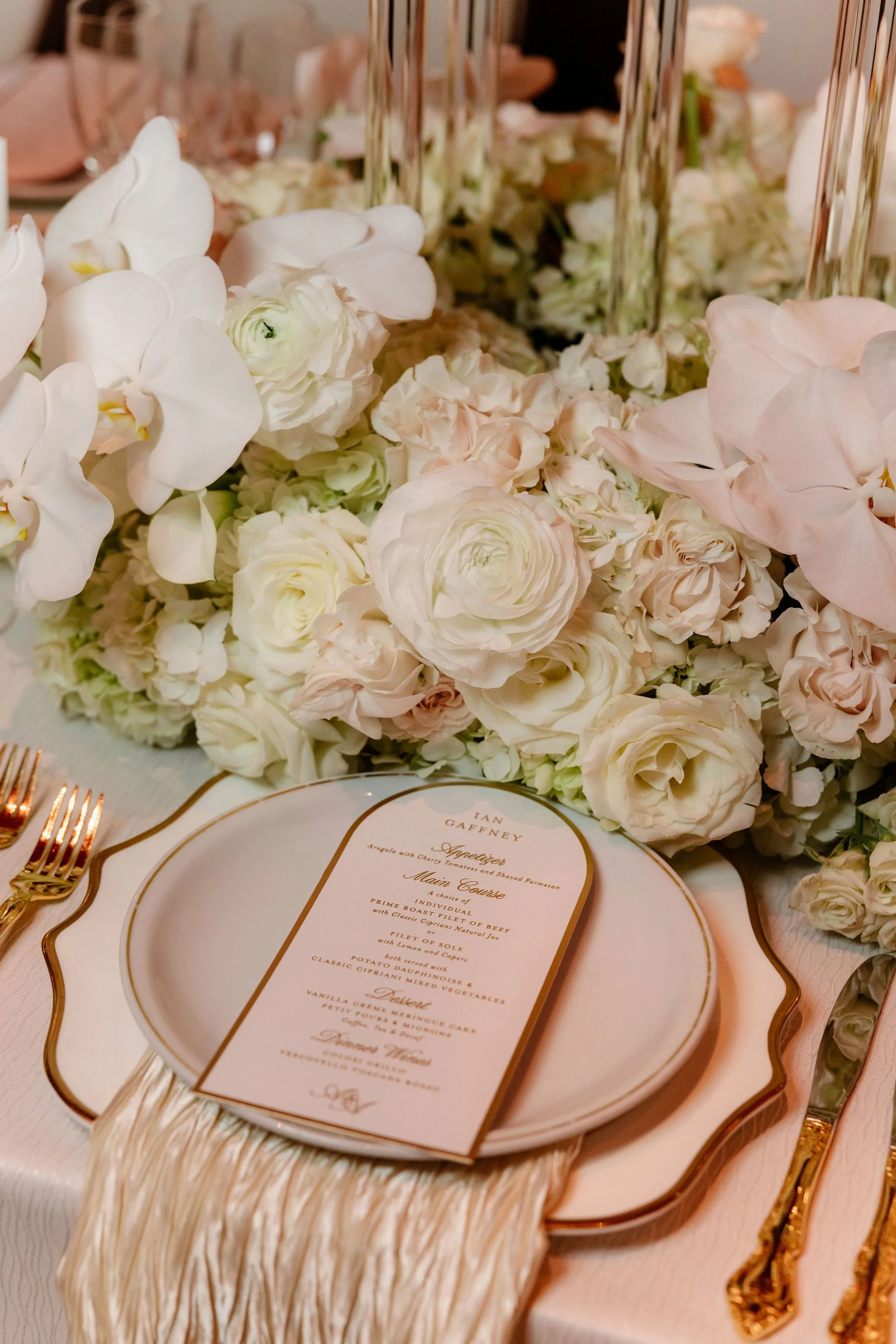 Close-up of a wedding reception table setting with a floral centerpiece, gold cutlery, a decorative menu card, and a textured beige table runner.